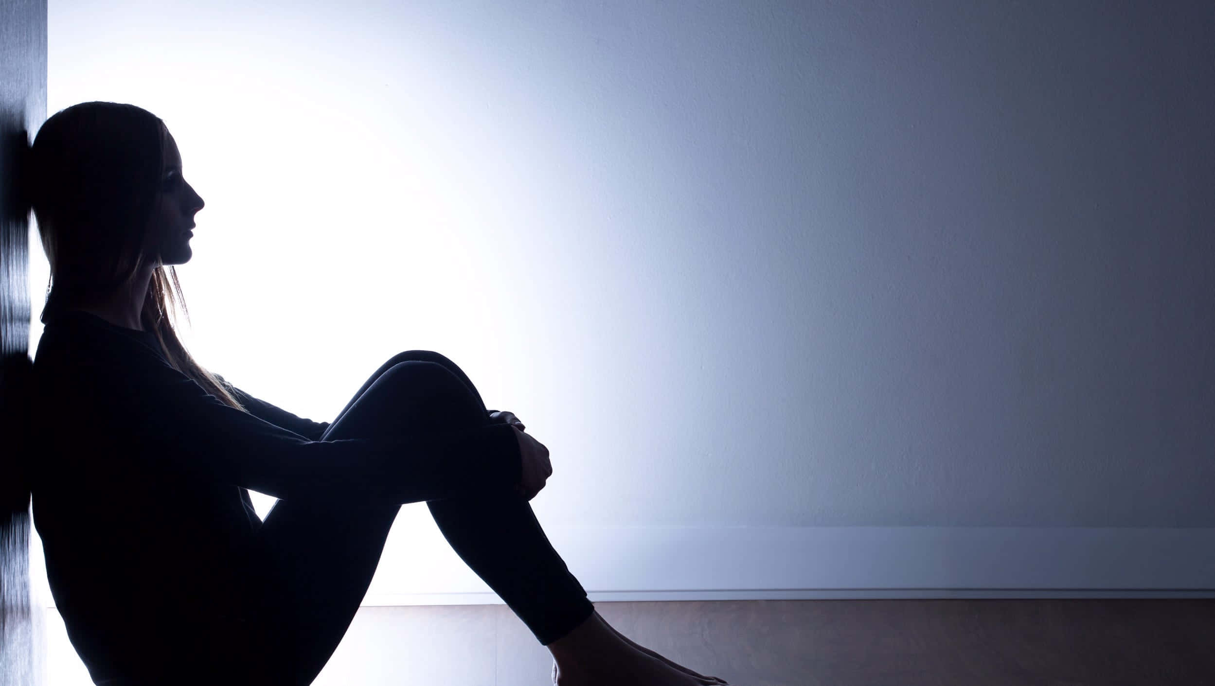 Depression Girl 桌布 壁紙 — 一個孤獨的女人坐在明亮的窗戶前，形成剪影，柔和的光線投射在樸素的牆壁上。