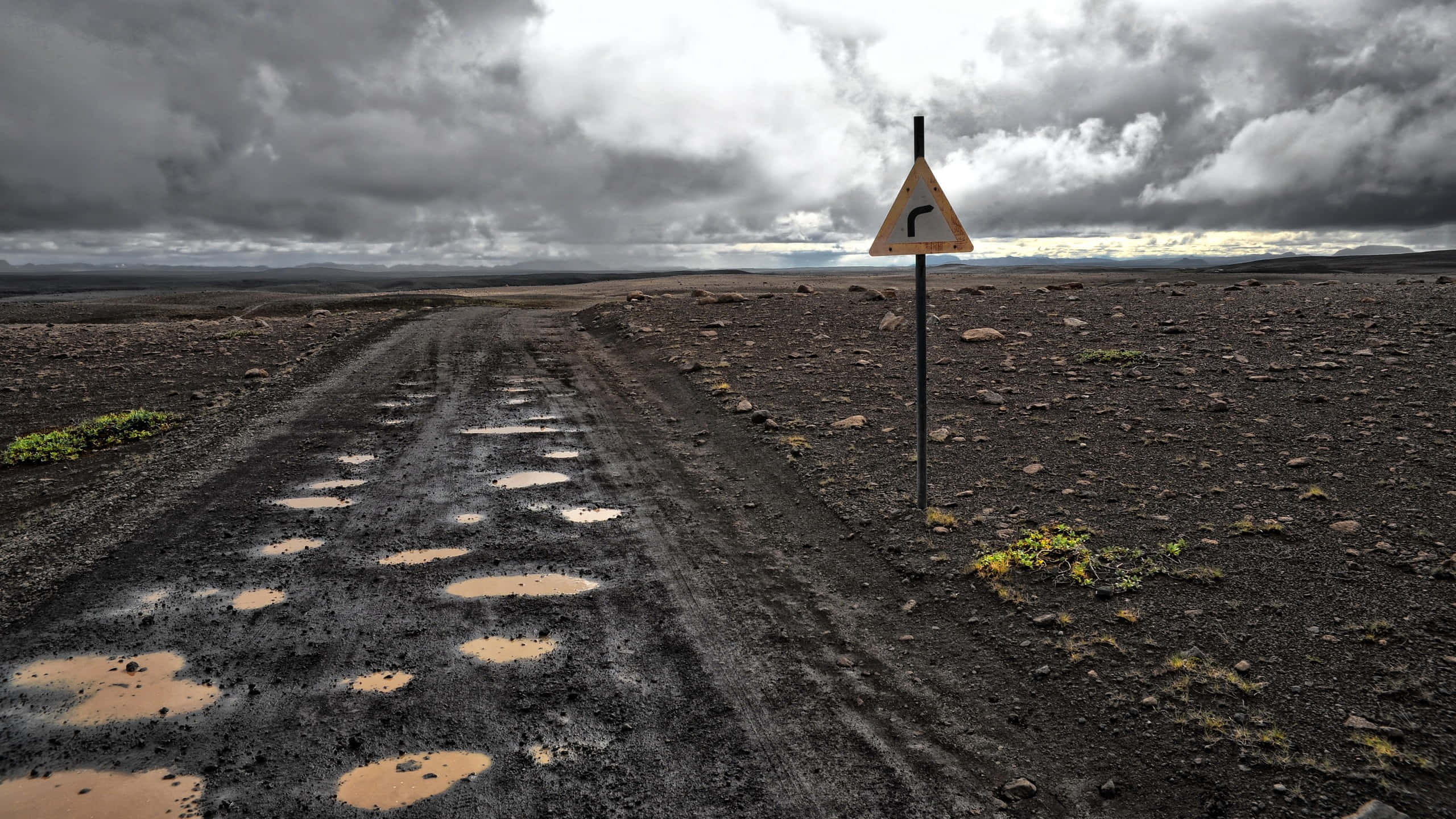 Road 桌布 壁紙 — 一個孤獨的彎曲路牌豎立在泥濘的碎石路旁，天空陰沉，背景是荒蕪的火山地貌。