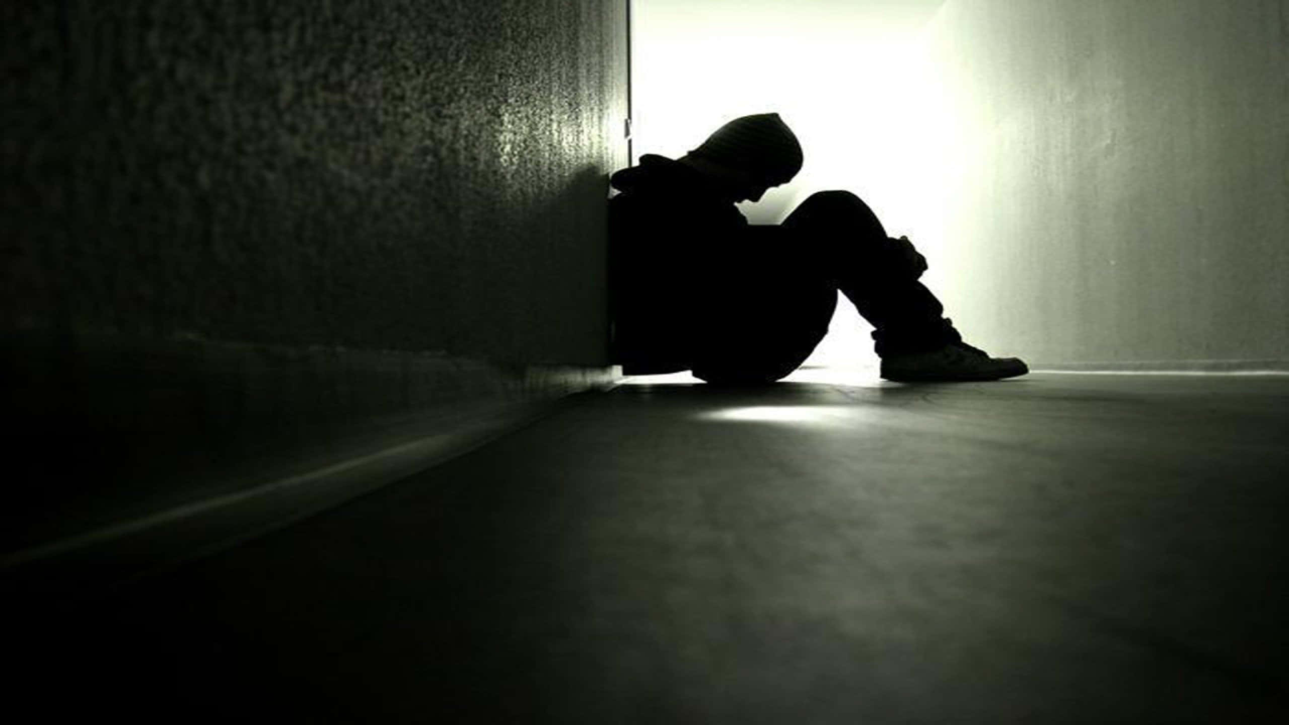 Alone 桌布 壁紙 — 一個孤獨的戴帽人影蜷縮在走廊角落的陰影中，沐浴在強烈的背光下。