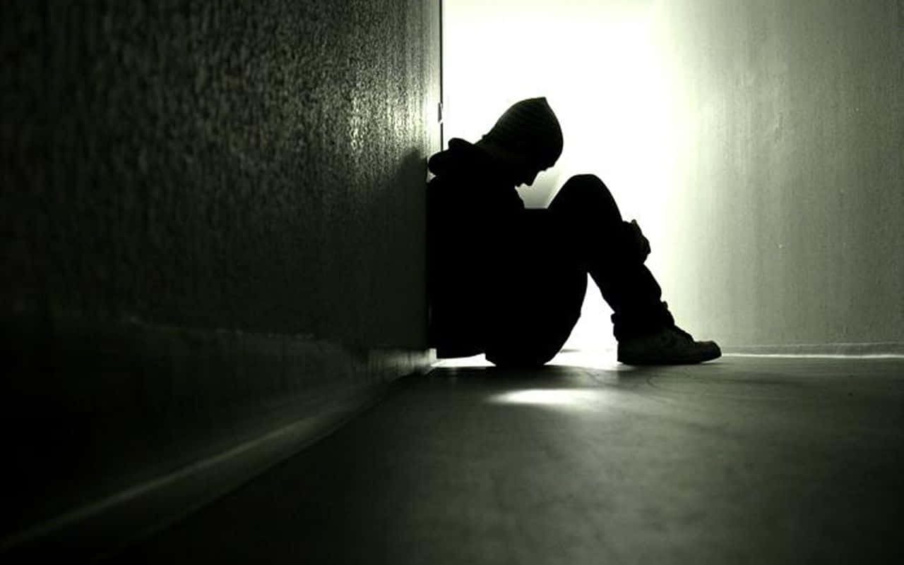 Sad Person 桌布 壁紙 — 一個孤獨的戴帽人物蜷縮在陰暗的角落，被強烈的背光照亮，在紋理牆壁上投射出長長的剪影。