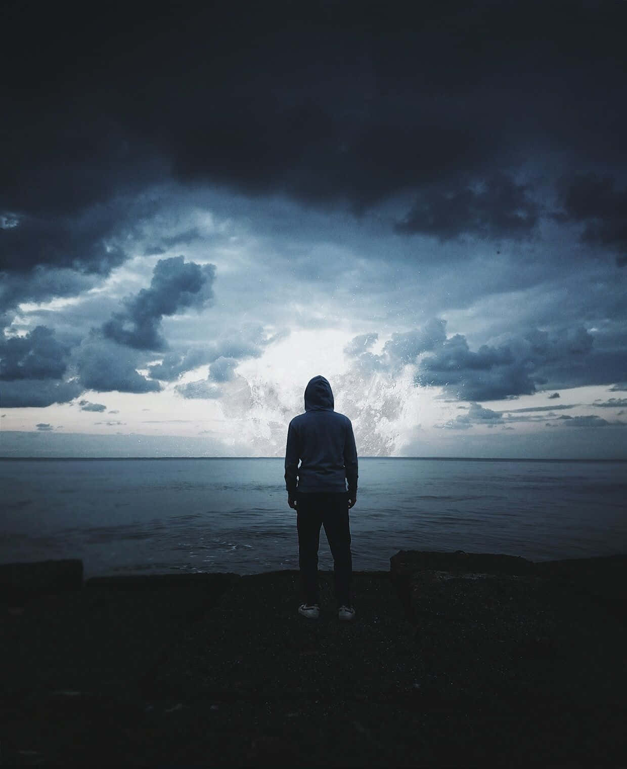 Hoodie 桌布 壁紙 — 一個孤獨的戴帽人站在岩石海岸上，面對廣闊的海洋，天空戲劇性地風起雲湧，雲層中有一道明亮的裂縫。