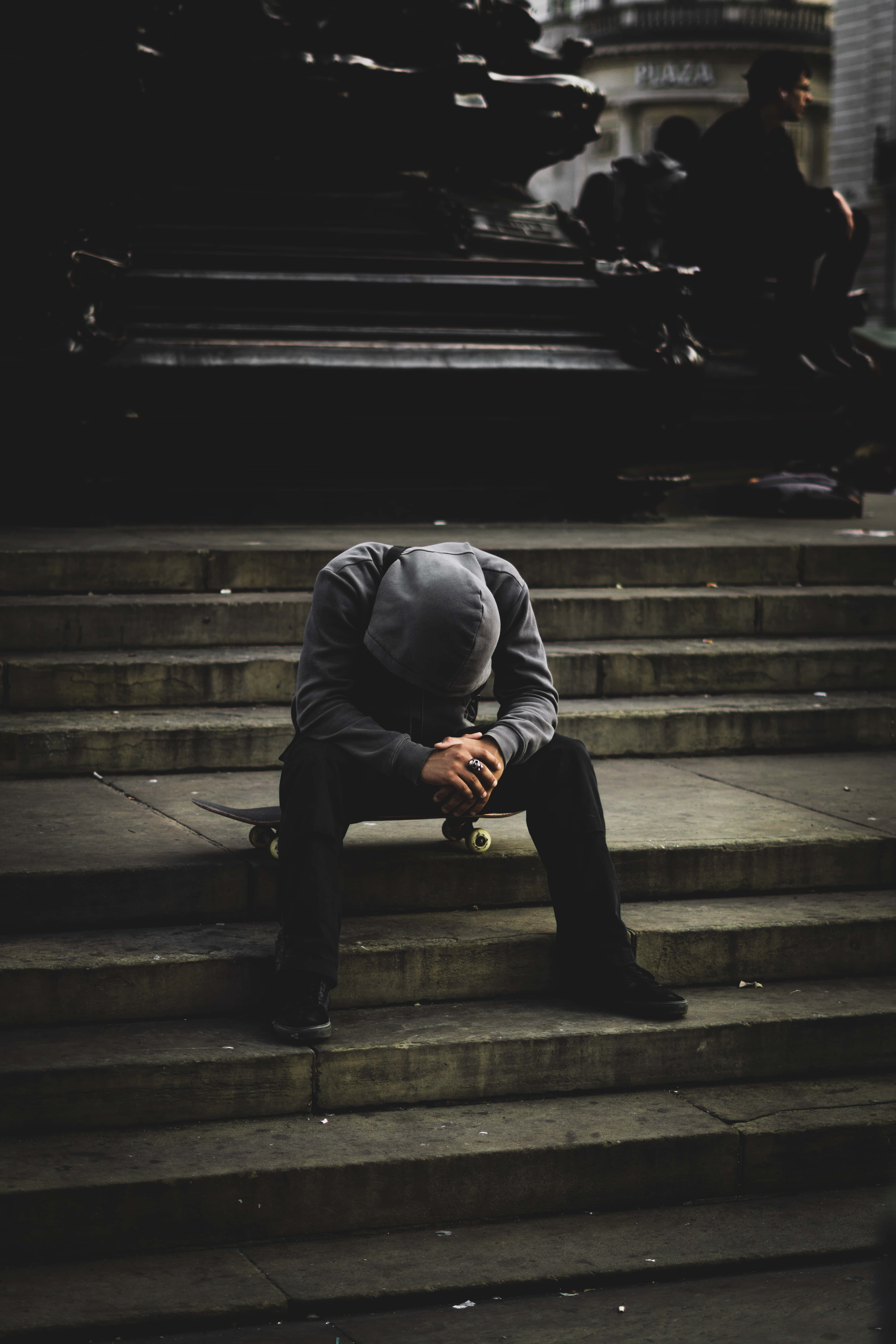 Alone 桌布 壁紙 — 一個孤獨的戴帽人蜷縮在石階上，拿著滑板，身處燈光昏暗的城市廣場。
