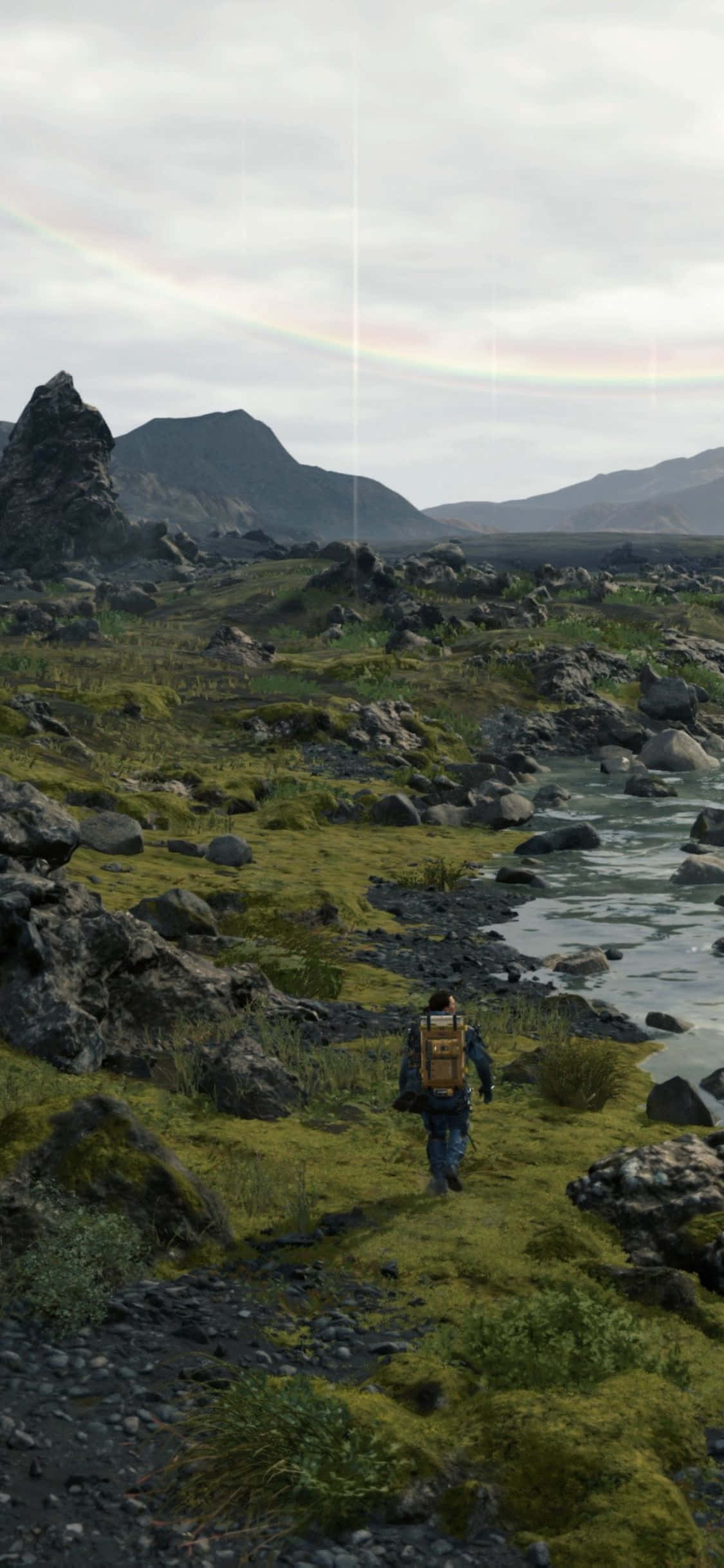 iPhone Xs Death Stranding 桌布 壁紙 — 一個孤獨的揹著背包的徒步旅行者走過一個長滿苔蘚的岩石山谷，一道淡淡的彩虹拱在遠山之上。