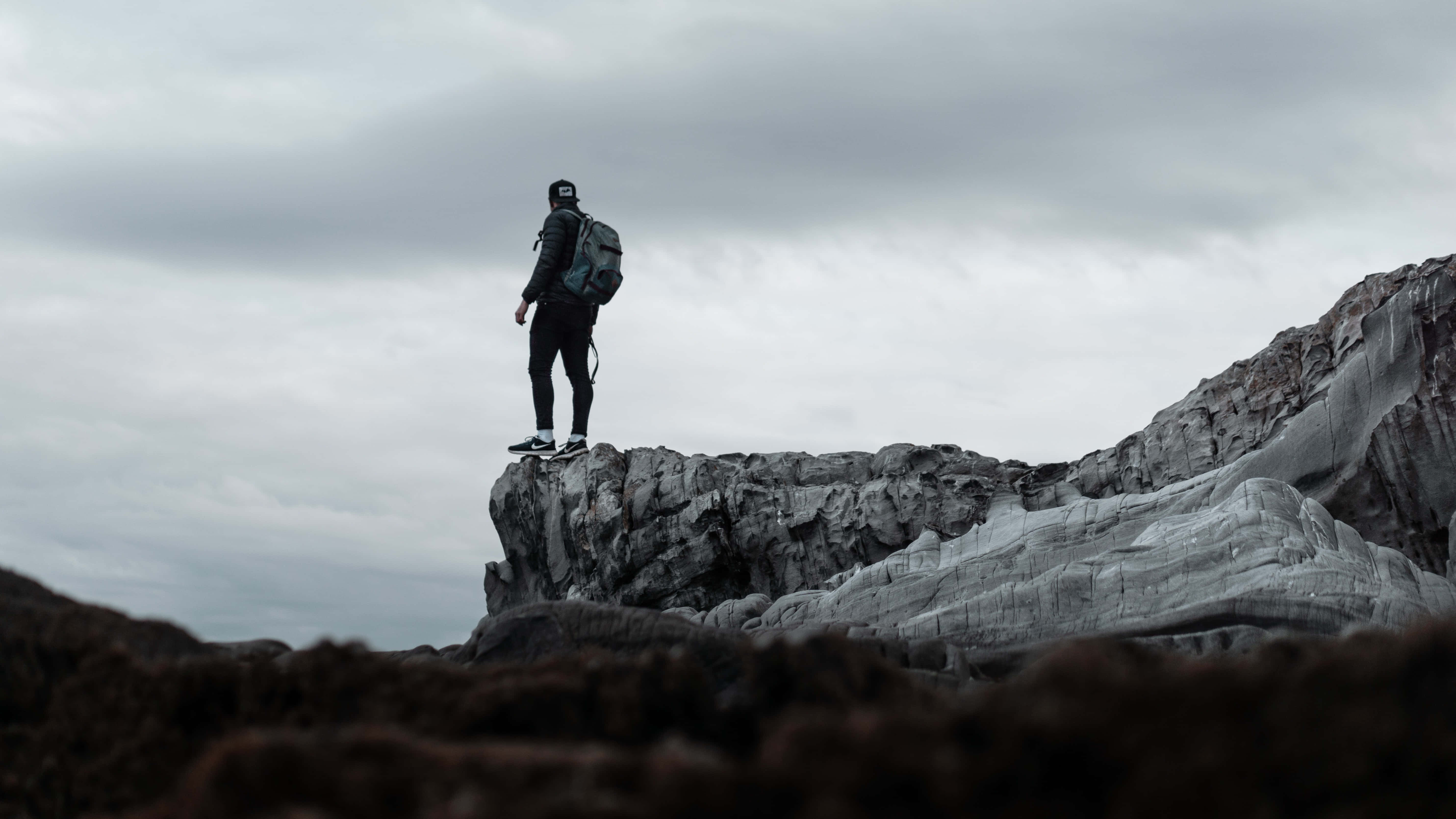 Climbing 桌布 壁紙 — 一個孤獨的揹著背包的遠足者站在崎嶇的懸崖邊緣，天空多雲。