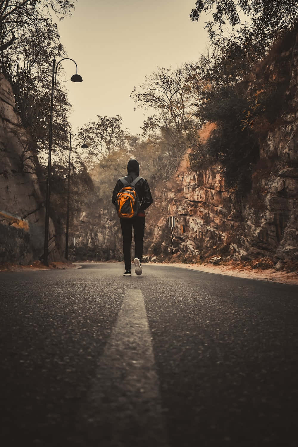 Walking 桌布 壁紙 — 一個孤獨的旅行者背著橙色背包，沿著蜿蜒的山路行走，兩旁是岩石峭壁和稀疏的樹木，天空朦朧。