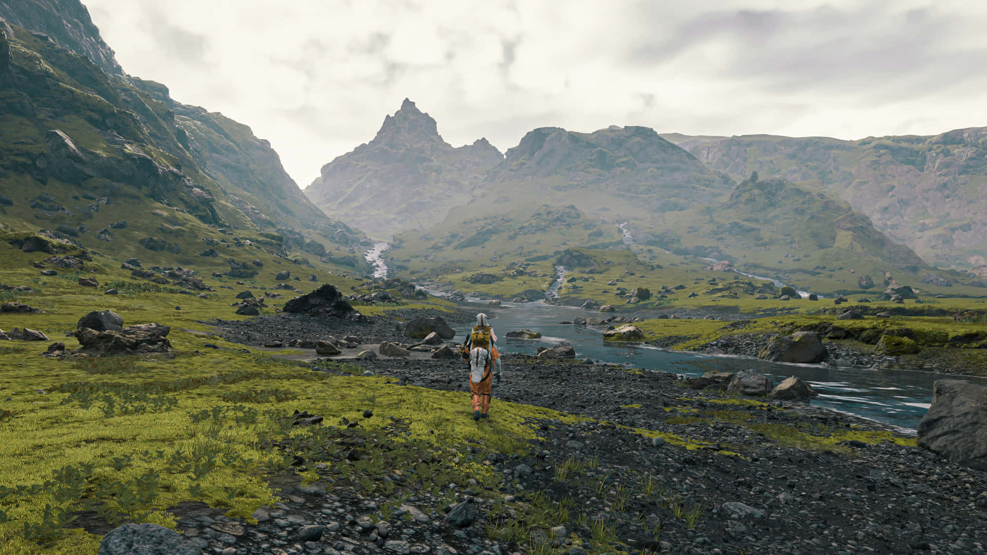 Land Scape Death Stranding Wide 壁紙 — 一個孤獨的旅行者背著背包，沿著岩石河岸穿過廣闊的綠色山谷，兩旁是高聳崎嶇的山脈，天空陰沉。