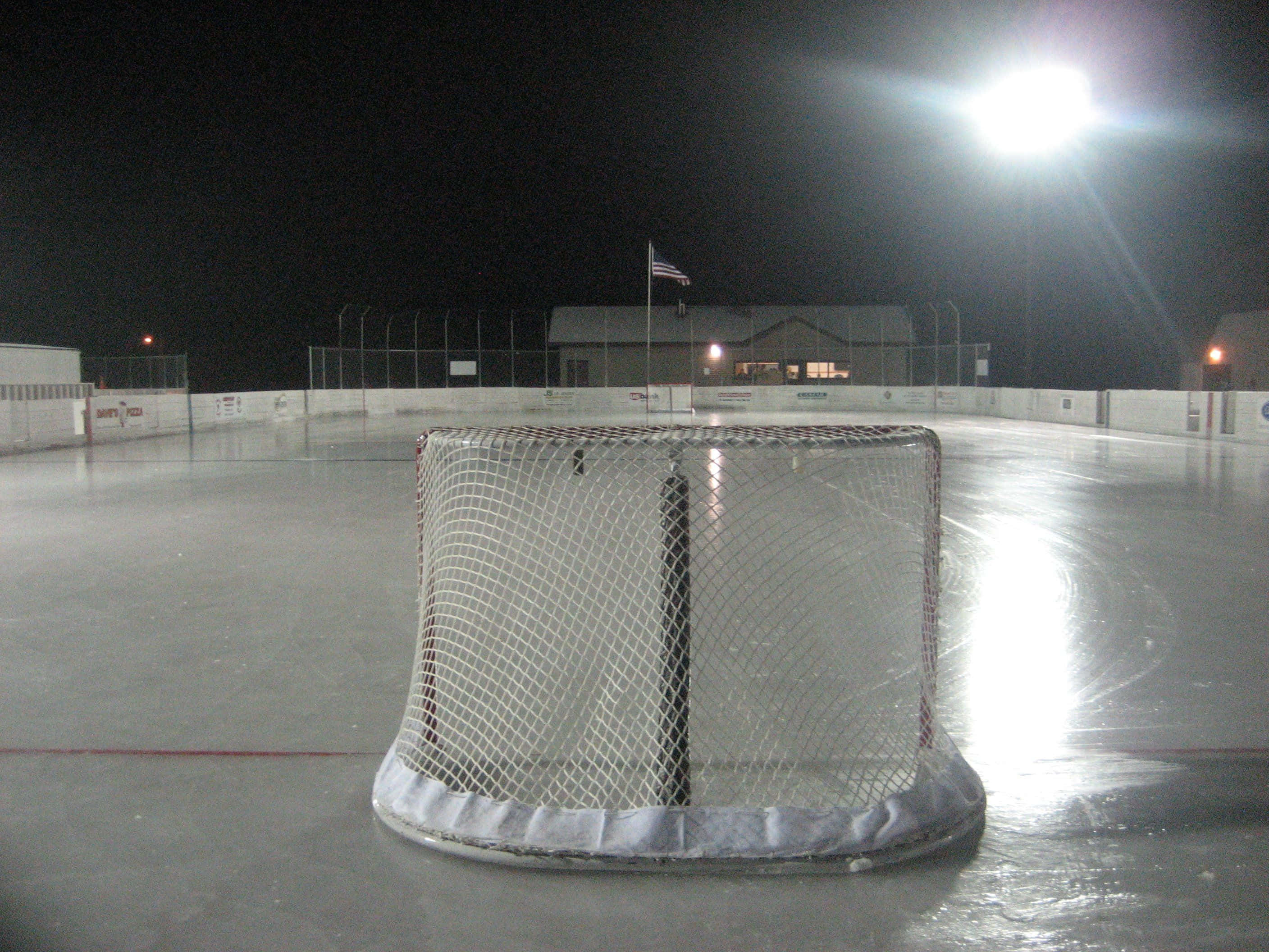 Ice Rink 桌布 壁紙 — 一個孤獨的曲棍球網在夜間空蕩蕩的溜冰場上，在明亮的聚光燈下發光，遠處有建築物和旗幟。