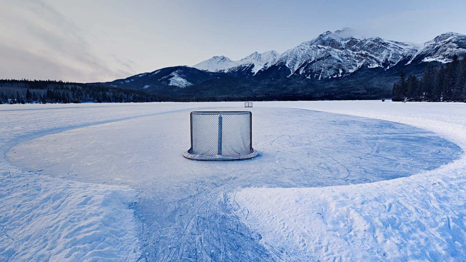 Ice Rink 桌布 壁紙 — 一個孤獨的曲棍球網立在冰凍的湖面上，周圍是白雪覆蓋的山脈，在蒼白的冬日天空下。