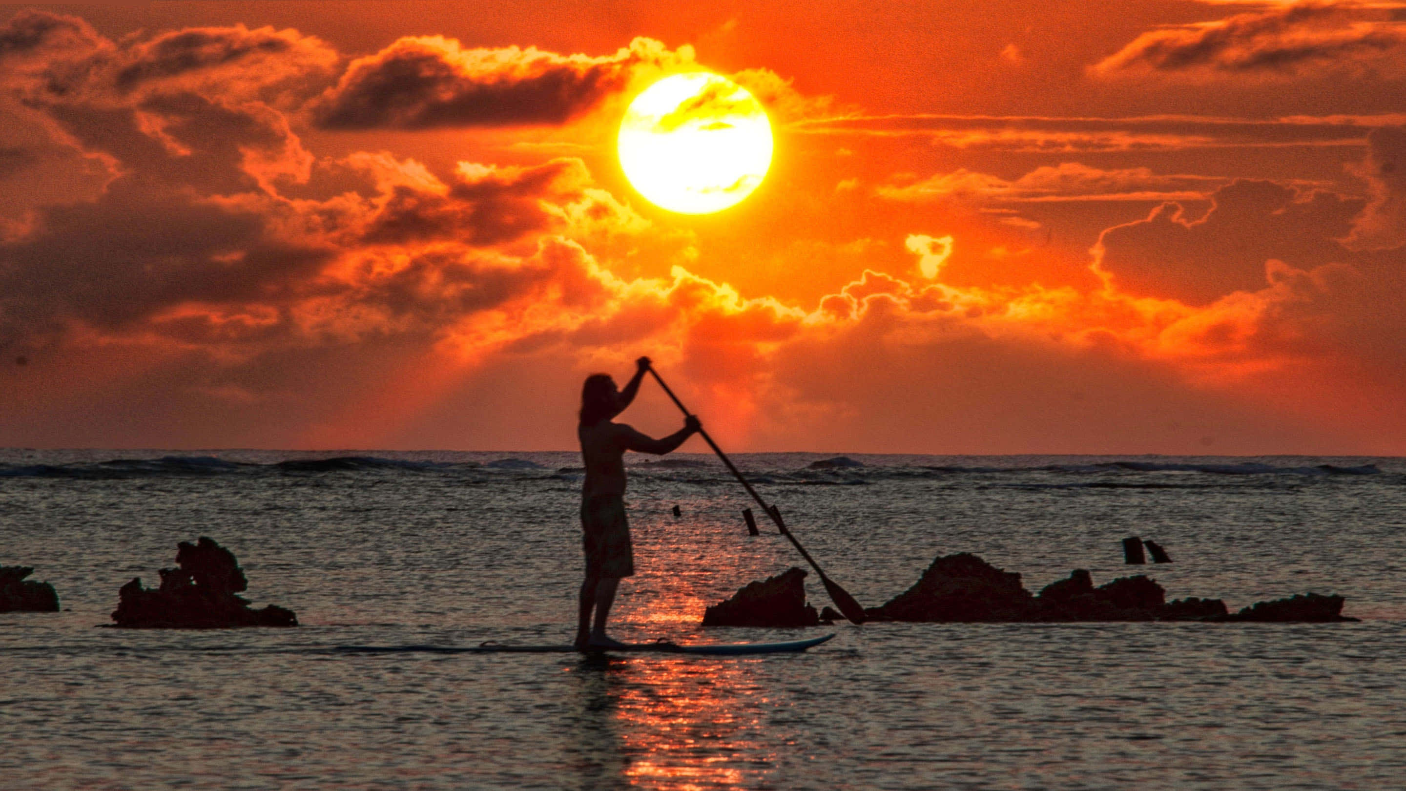 Paddleboarding 桌布 壁紙 — 一個孤獨的槳板衝浪者，在平靜的海面上，以火紅的橙色日落為背景，剪影中散佈著岩石。