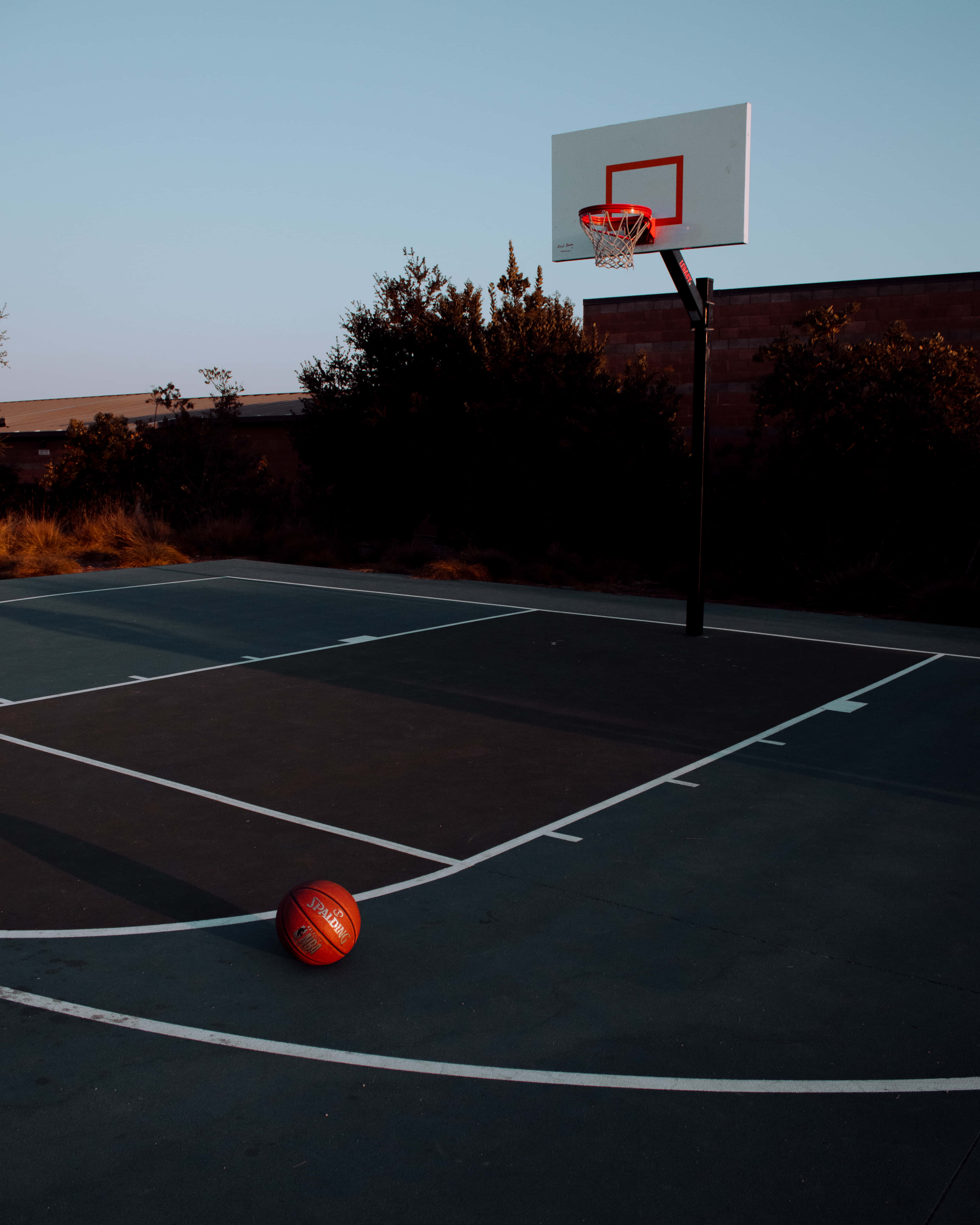 籃球 桌布 壁紙 — 一個孤獨的橙色籃球放在昏暗的戶外球場上，黃昏時分，籃框在漸暗的天空中呈現剪影。