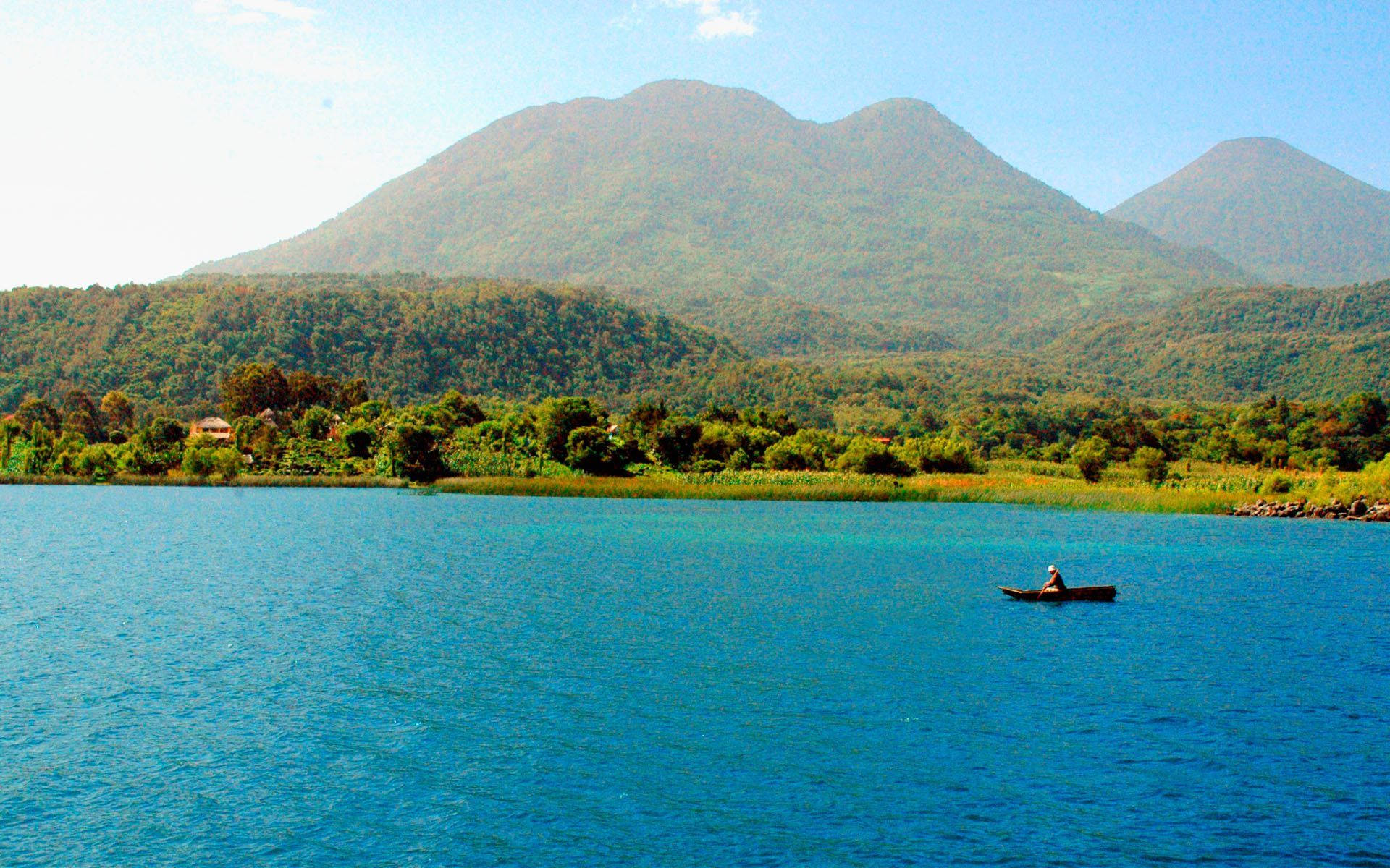 Guatemala 桌布 壁紙 — 一個孤獨的漁夫在小船上滑過充滿活力的藍色湖泊，背景是鬱鬱蔥蔥的綠色山脈在晴朗的天空下雄偉地升起。