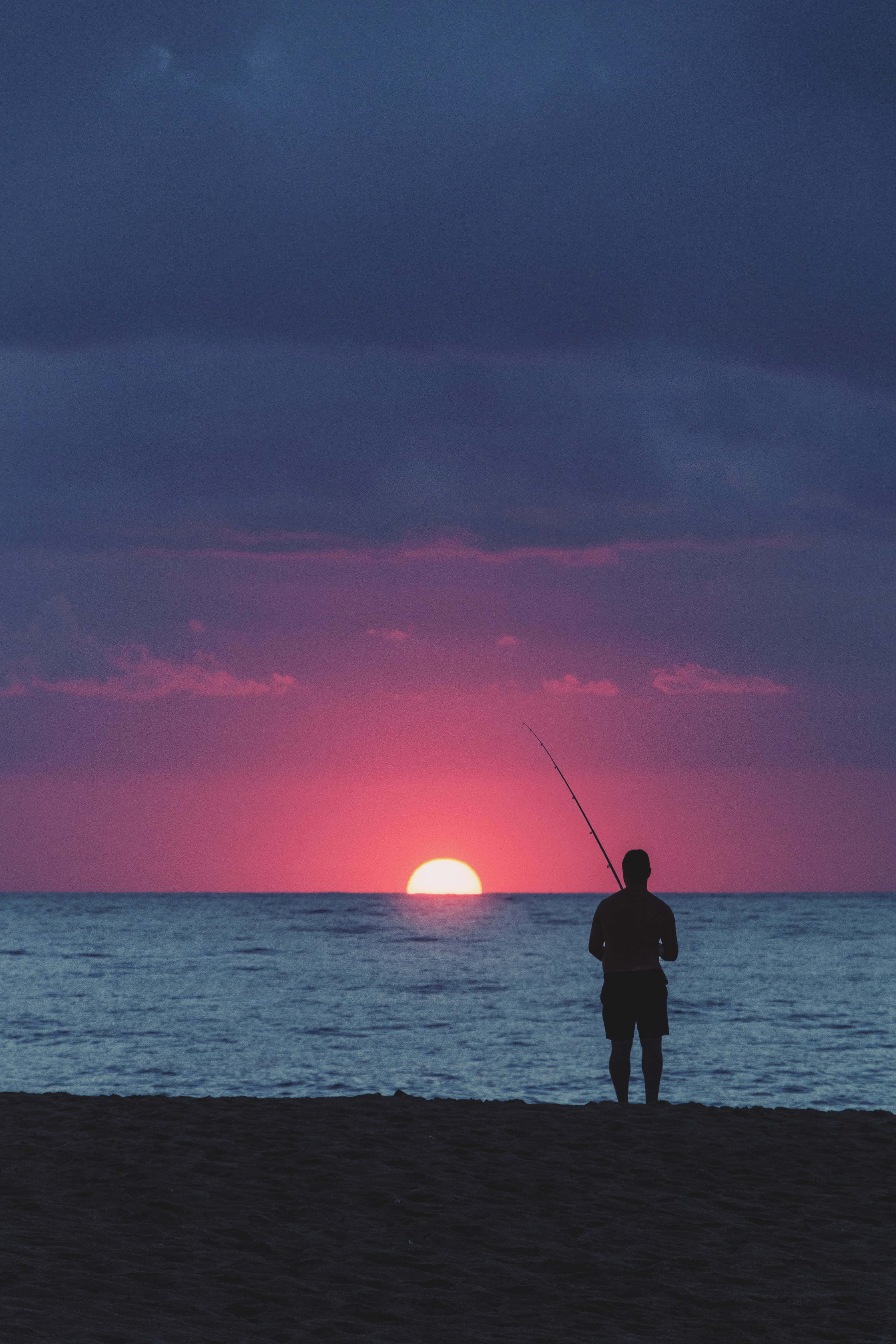 Fishing Phone 桌布 壁紙 — 一個孤獨的漁夫在海灘上以剪影姿態站立，在夕陽沉入火紅與深紫雲層之際拋出釣線。