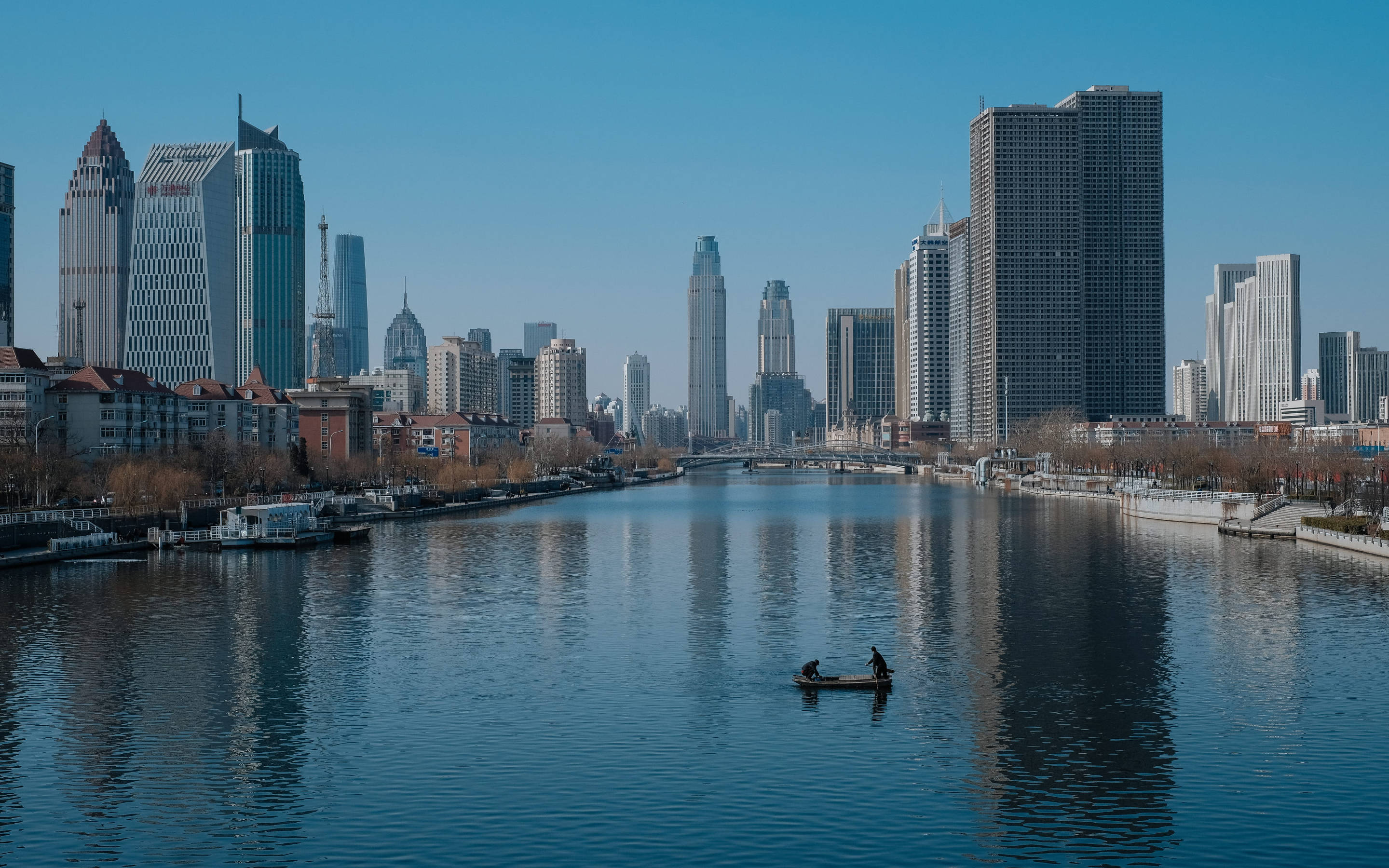 Tianjin 桌布 壁紙 — 一個孤獨的漁夫坐在小船裡，在平靜的河面上滑行，河水映照著晴朗藍天下高聳的現代摩天大樓。