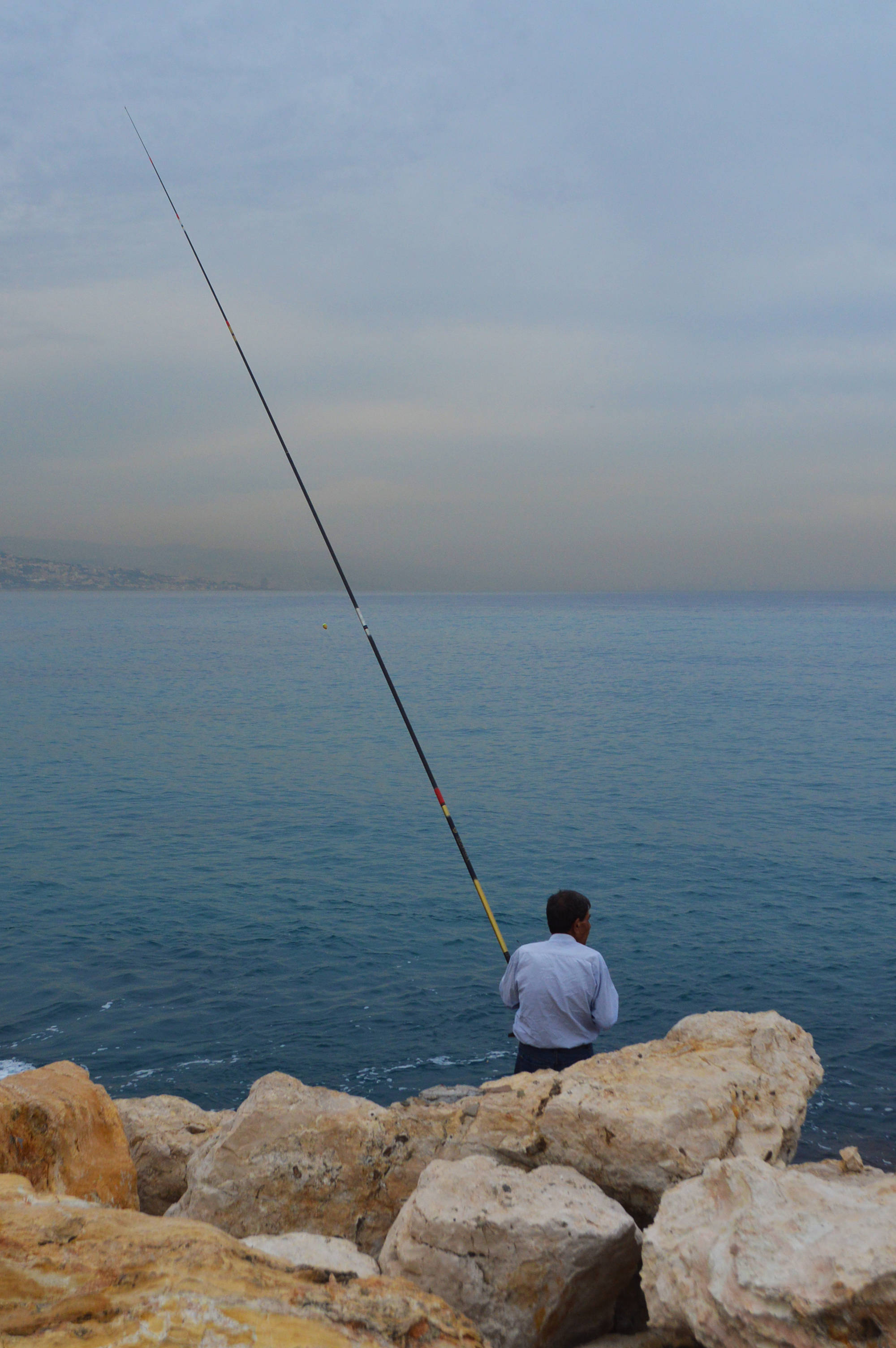 Lebanon 桌布 壁紙 — 一個孤獨的漁夫站在岩石峭壁上，在陰天之下將釣線拋入平靜的藍色大海。