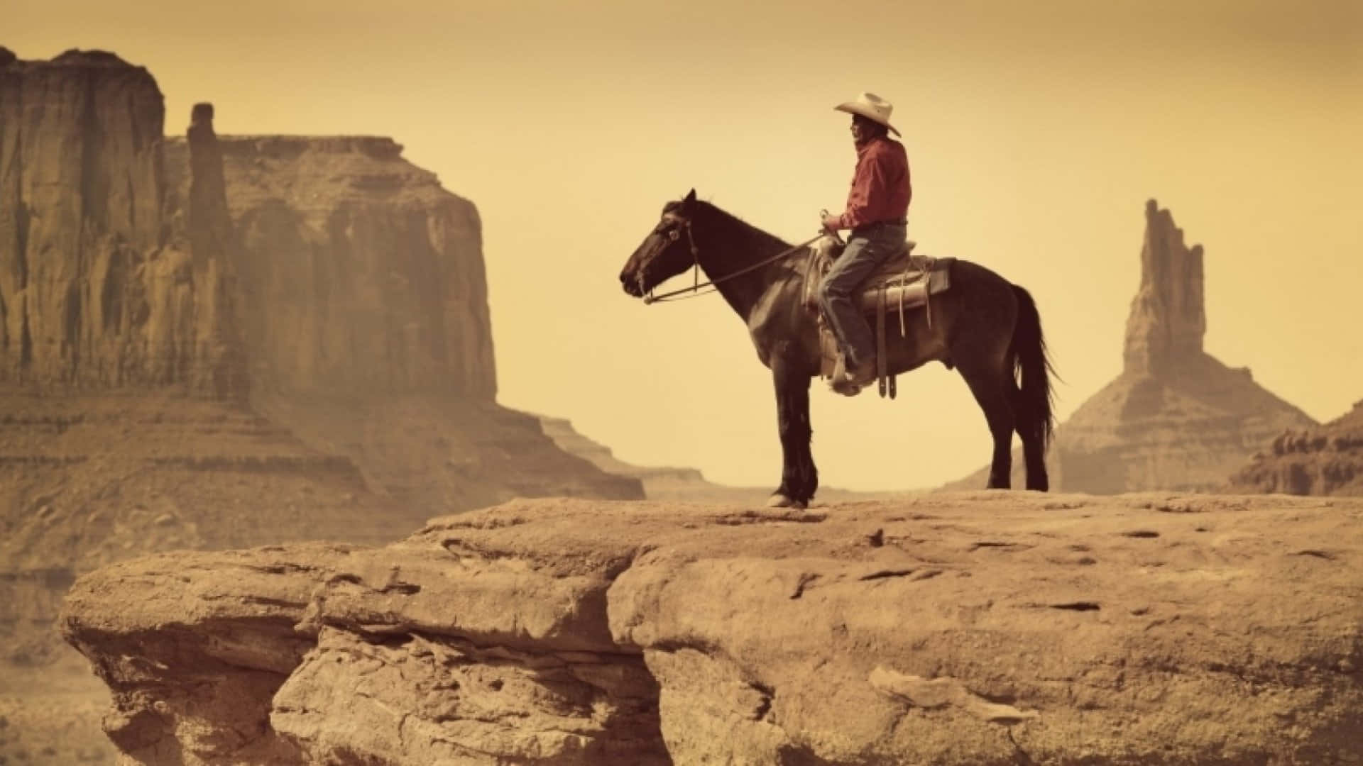 Old Western 桌布 壁紙 — 一個孤獨的牛仔，身穿紅衫、頭戴白帽，騎著黑馬站在岩石峭壁上，俯瞰廣闊的沙漠峽谷，天空籠罩著朦朧的金色光芒。