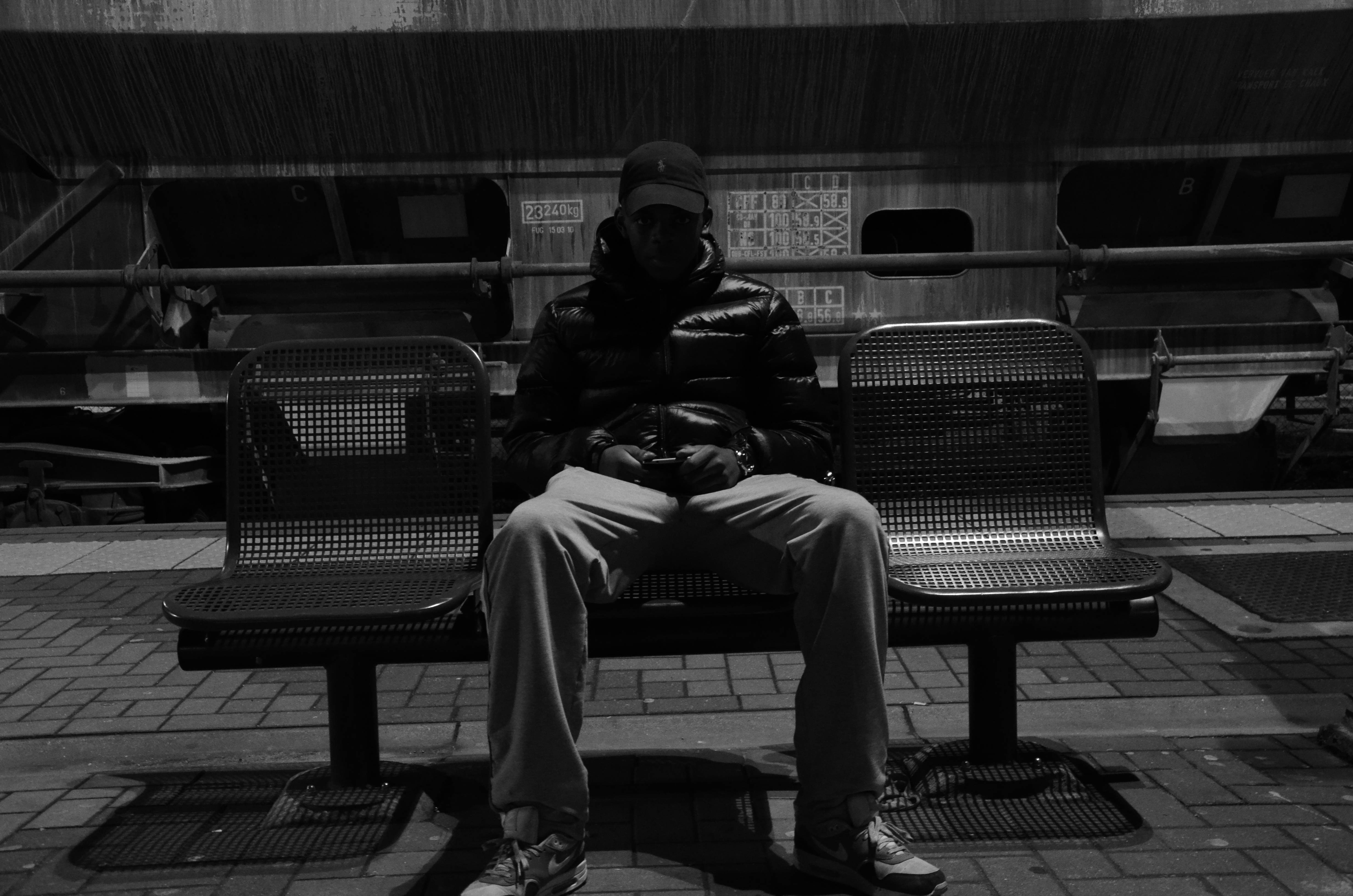 tired 桌布 壁紙 — 一個孤獨的男人穿著羽絨外套，坐在火車站月台的金屬長椅上，黑白畫面。