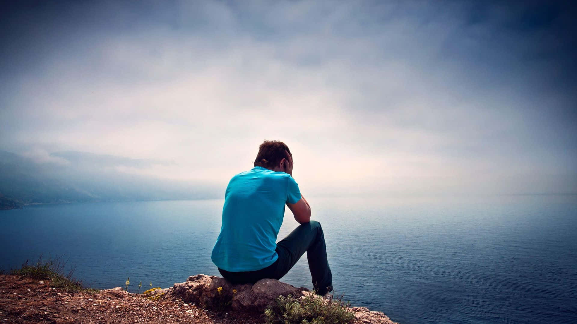 Alone 桌布 壁紙 — 一個孤獨的男人穿著藍色襯衫坐在岩石懸崖邊緣，凝視著多雲天空下廣闊的霧海。