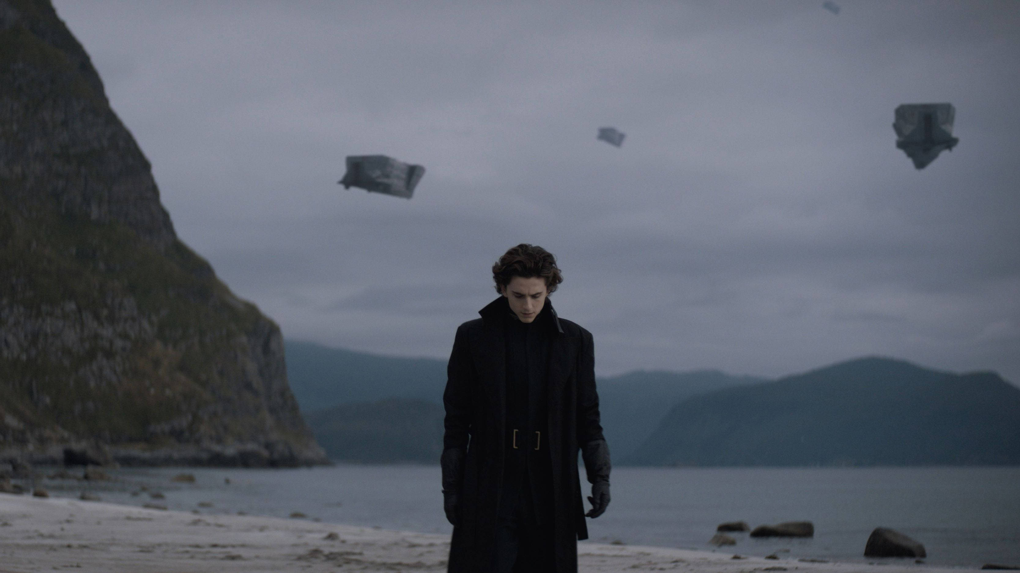 Dune Movie 桌布 壁紙 — 一個孤獨的男人穿著黑色外套站在霧氣瀰漫的海岸沙灘上，漂浮的石頭巨石在陰天中漂流。