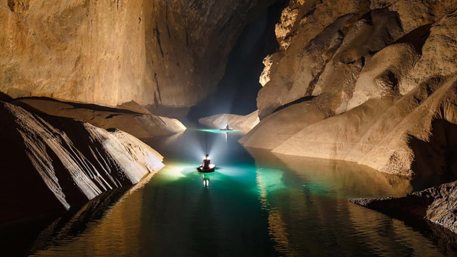 Cave 桌布 壁紙 — 一個孤獨的皮划艇運動員滑行穿過一個廣闊、昏暗的洞穴，發光的藍綠色水面反射著溫暖的岩壁。