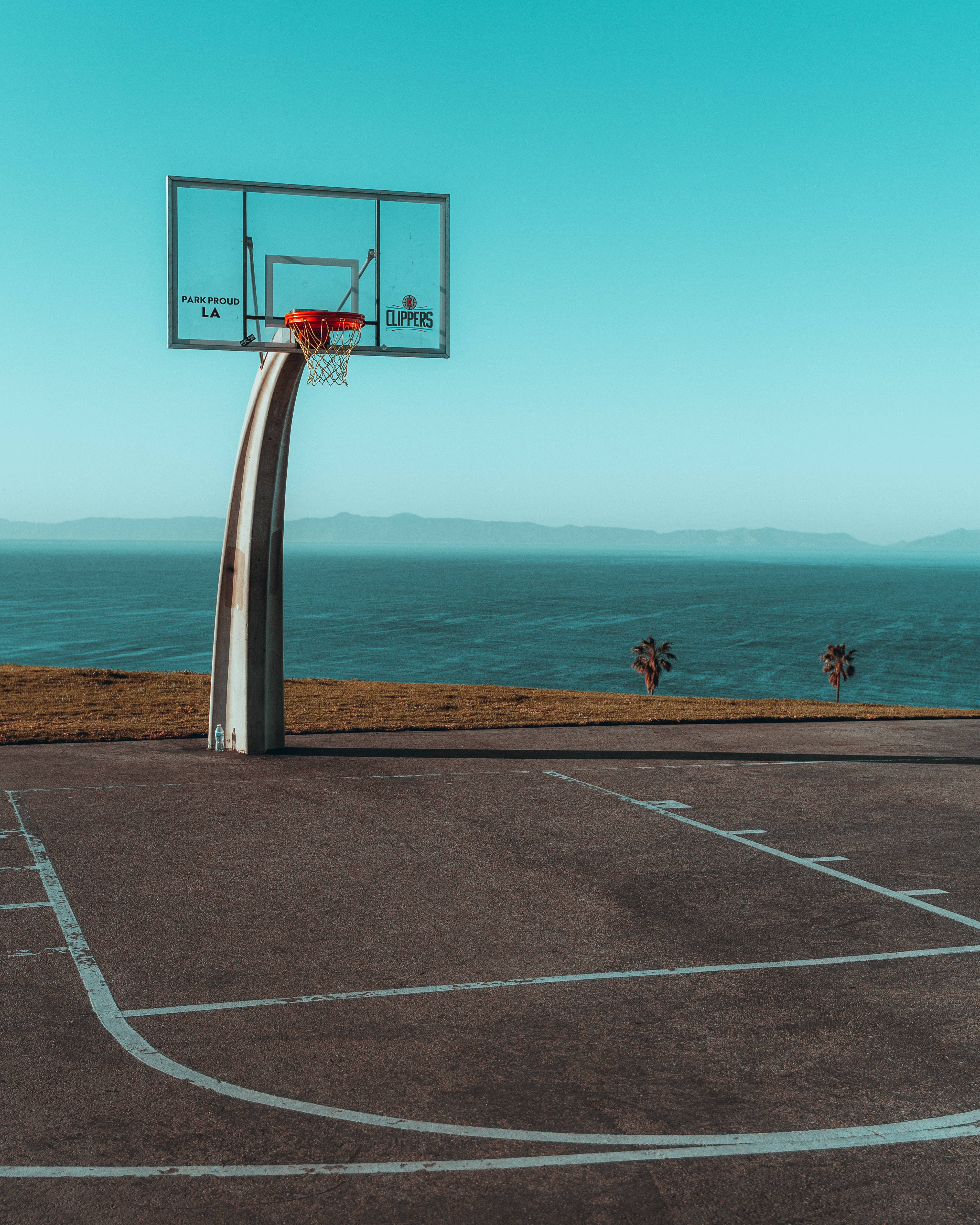Playground 桌布 壁紙 — 一個孤獨的籃球框矗立在沿海球場上，俯瞰著海洋和遠處的山脈，天空清澈湛藍。