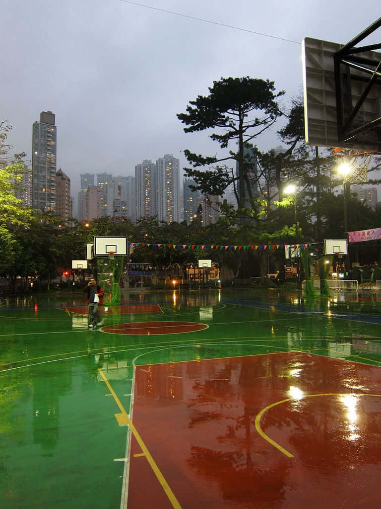 jump 桌布 壁紙 — 一個孤獨的籃球運動員在雨濕的球場上運球，城市燈光下，高聳的摩天大樓在霧濛濛的背景中若隱若現。