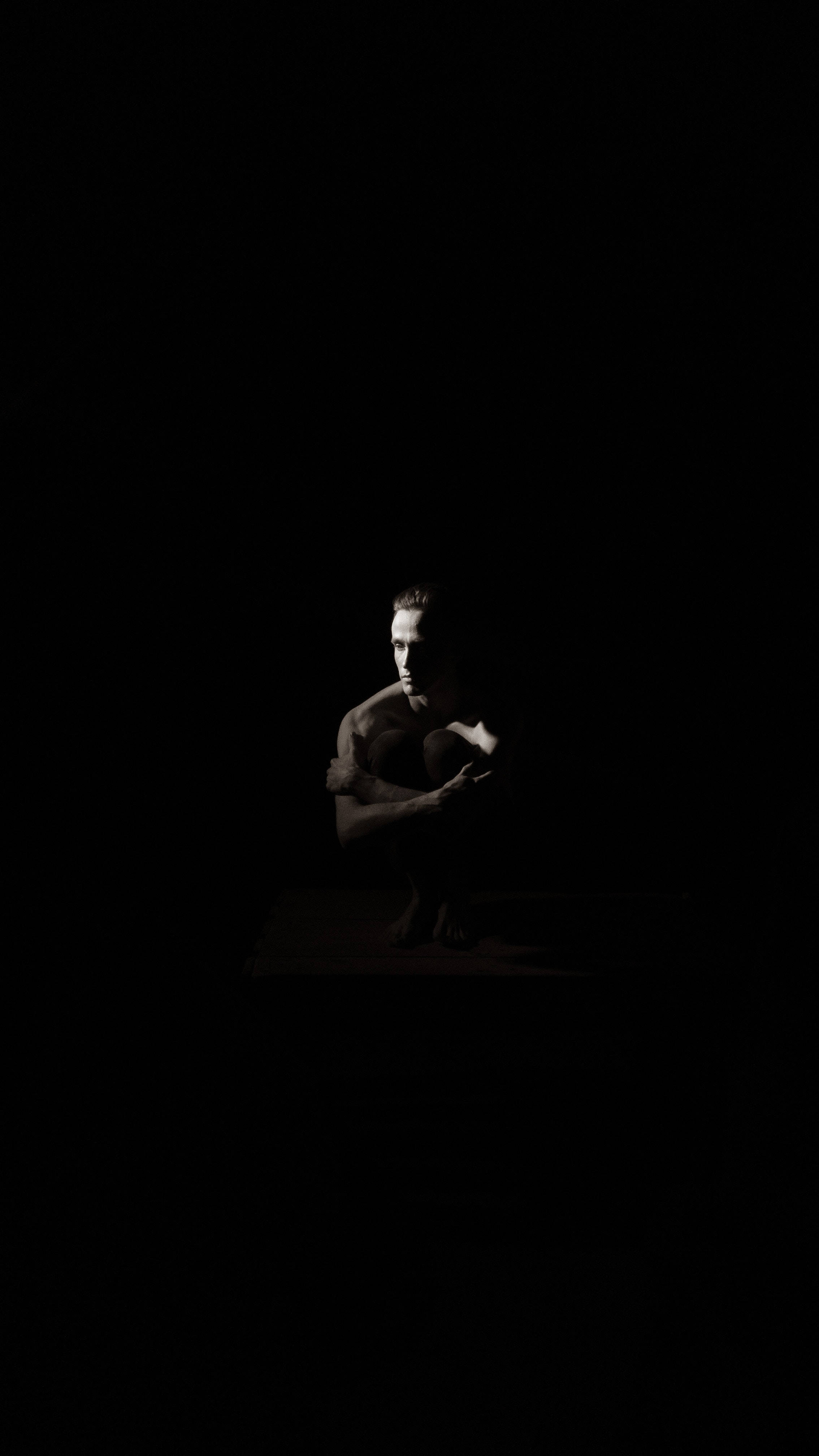 黑白 桌布 壁紙 — 一個孤獨的肌肉男坐在深沉的陰影中，戲劇性的聚光燈照亮他的臉部和軀幹，背景是漆黑一片。