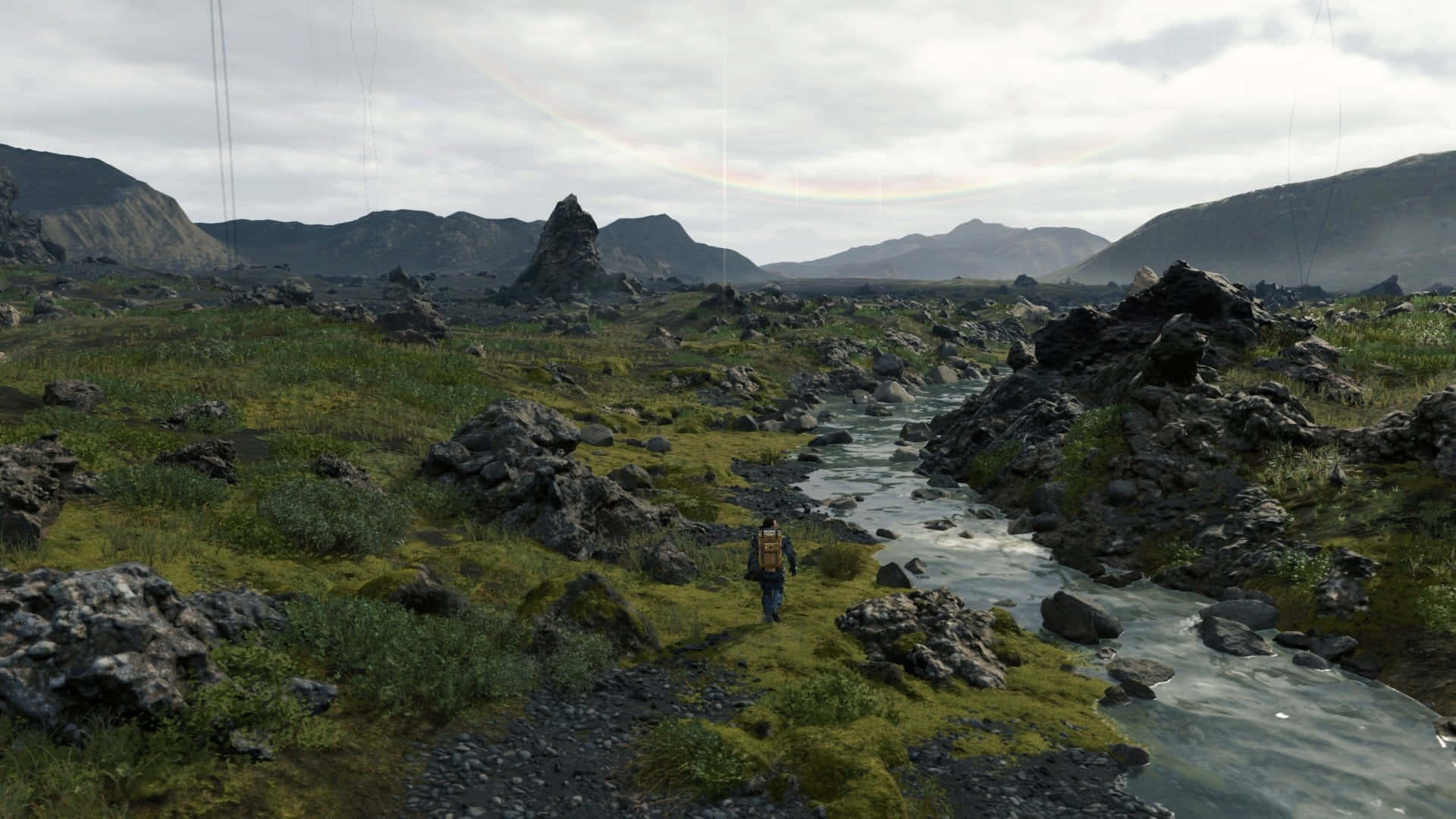 HD Death Stranding 桌布 壁紙 — 一個孤獨的背著背包的徒步旅行者，走過一個長滿苔蘚的岩石山谷，山谷中有一條蜿蜒的小溪，天空多雲，有一道淡淡的彩虹。