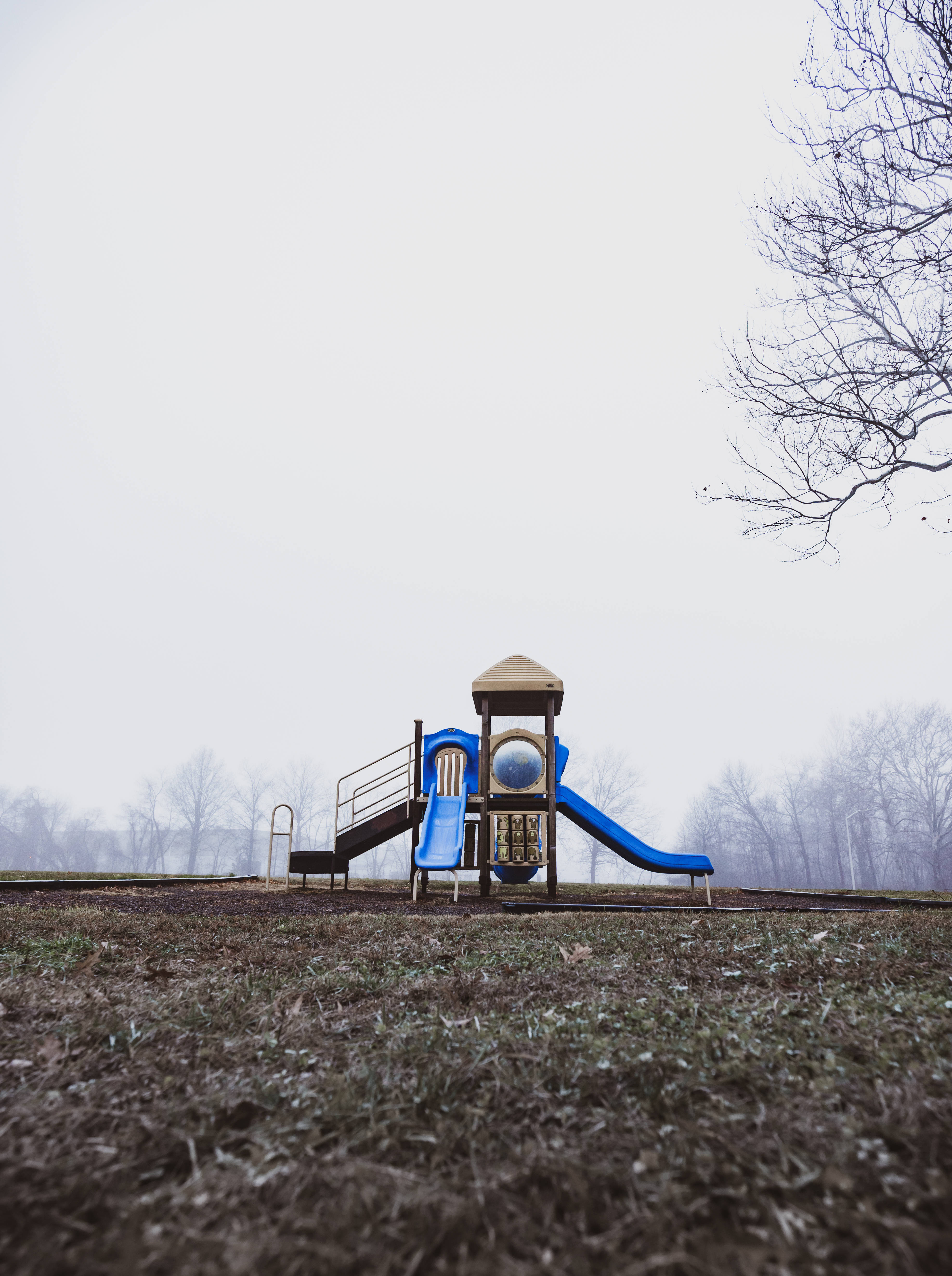 Playground 桌布 壁紙 — 一個孤獨的藍色溜滑梯靜靜地矗立在霧氣瀰漫的田野中，周圍是光禿禿的樹木，天空陰沉。