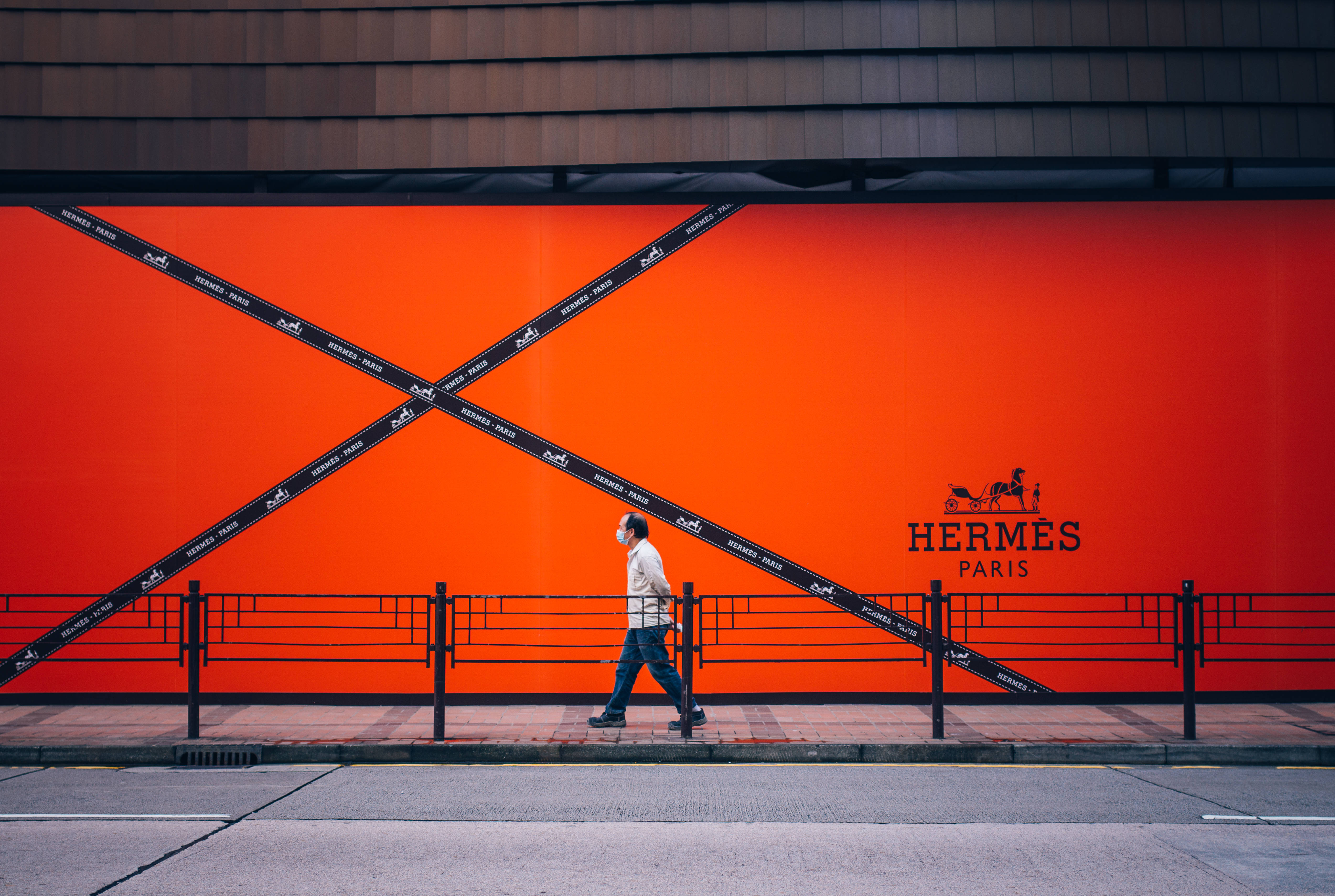 Hermes 桌布 壁紙 — 一個孤獨的行人走過一個現代城市環境中，被黑色斜膠帶交叉的鮮橙色愛馬仕廣告看板。