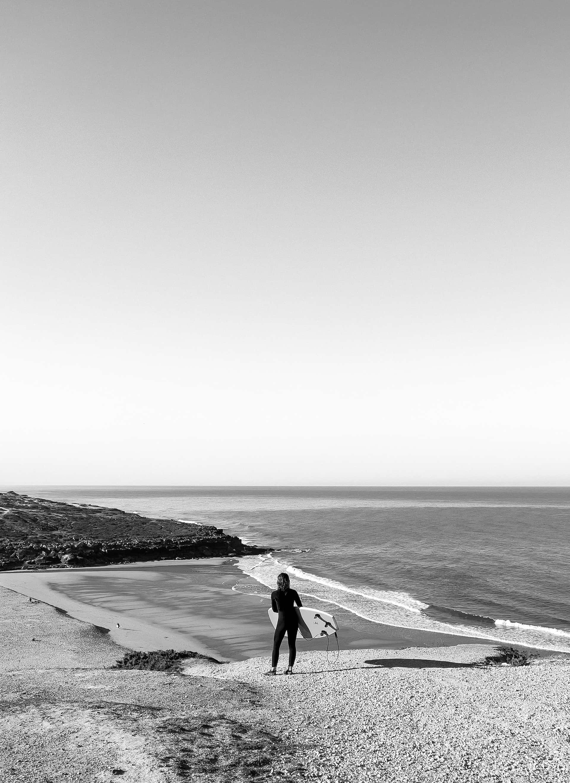 Black And White Beach 桌布 壁紙 — 一個孤獨的衝浪者穿著潛水衣站在卵石灘上，手持衝浪板，面向廣闊晴朗天空下平靜的海洋。