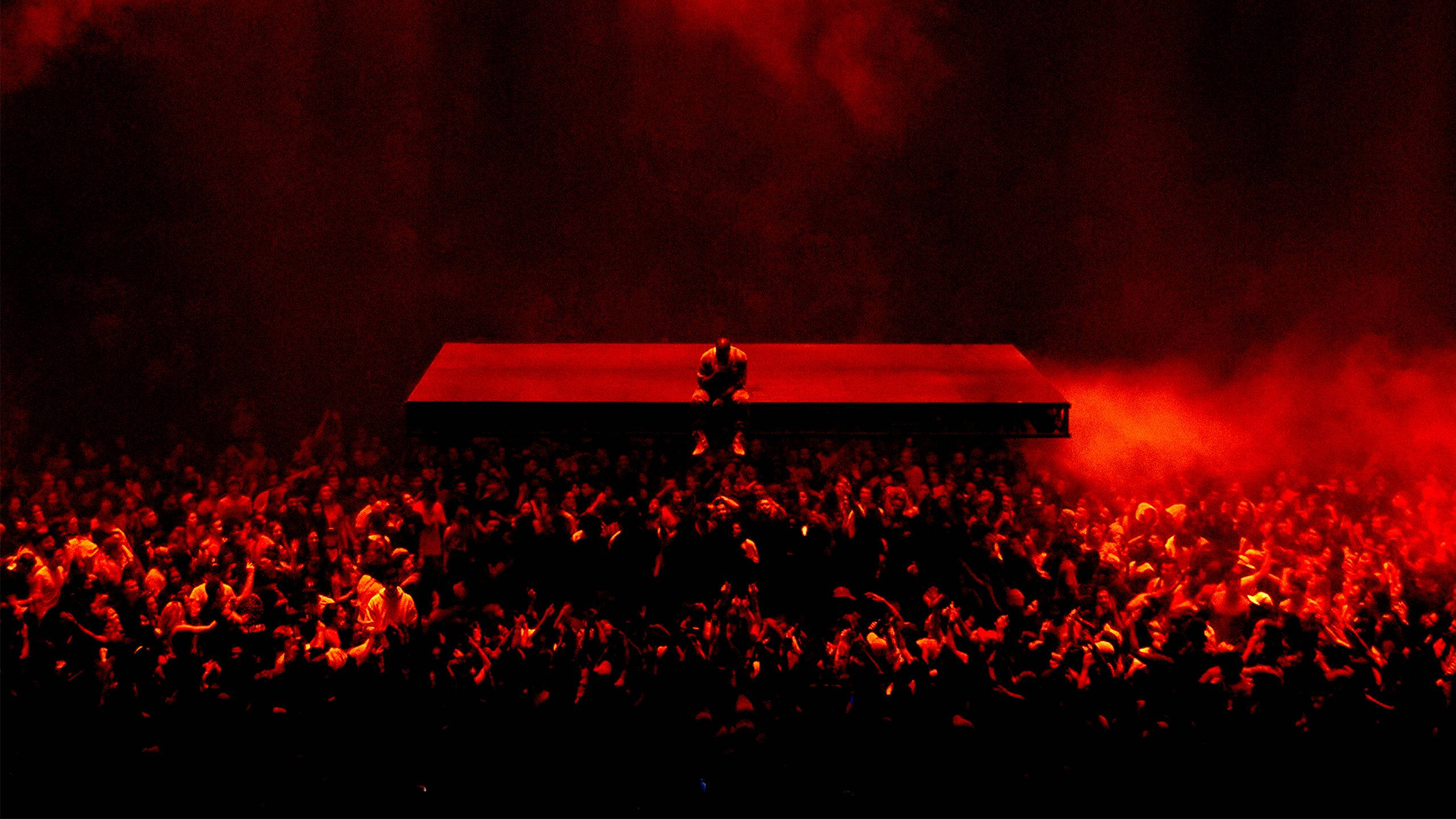 kanye west saint pablo 桌布 壁紙 — 一個孤獨的表演者剪影在一個被紅光籠罩的舞台上，下方是一片在煙霧中迷失的歡呼音樂會觀眾海洋。