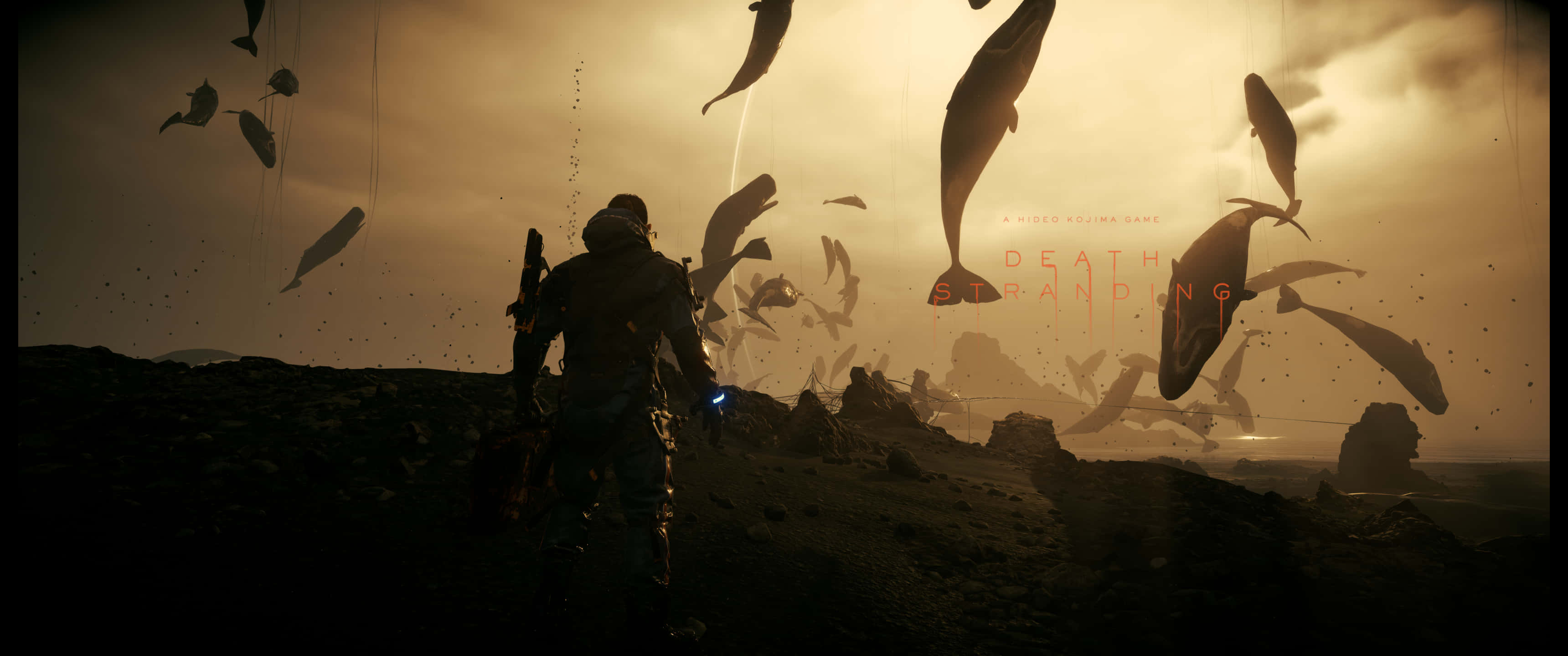 3440x1440p Death Stranding 桌布 壁紙 — 一個孤獨的裝甲人物站在貧瘠的景觀上，而巨大的鯨魚狀生物漂浮在朦朧的金色天空中。