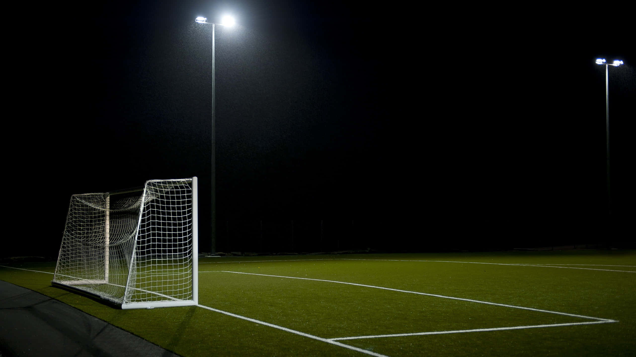 sport 桌布 壁紙 — 一個孤獨的足球龍門在夜間空蕩蕩的人造草地球場上，在明亮的泛光燈下發光。
