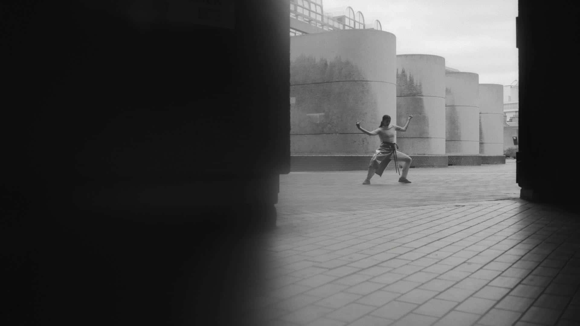 黑白 桌布 壁紙 — 一個孤獨的身影以動態武術姿勢站在黑暗拱門之間，背景是陰天下的巨大圓柱形儲罐。