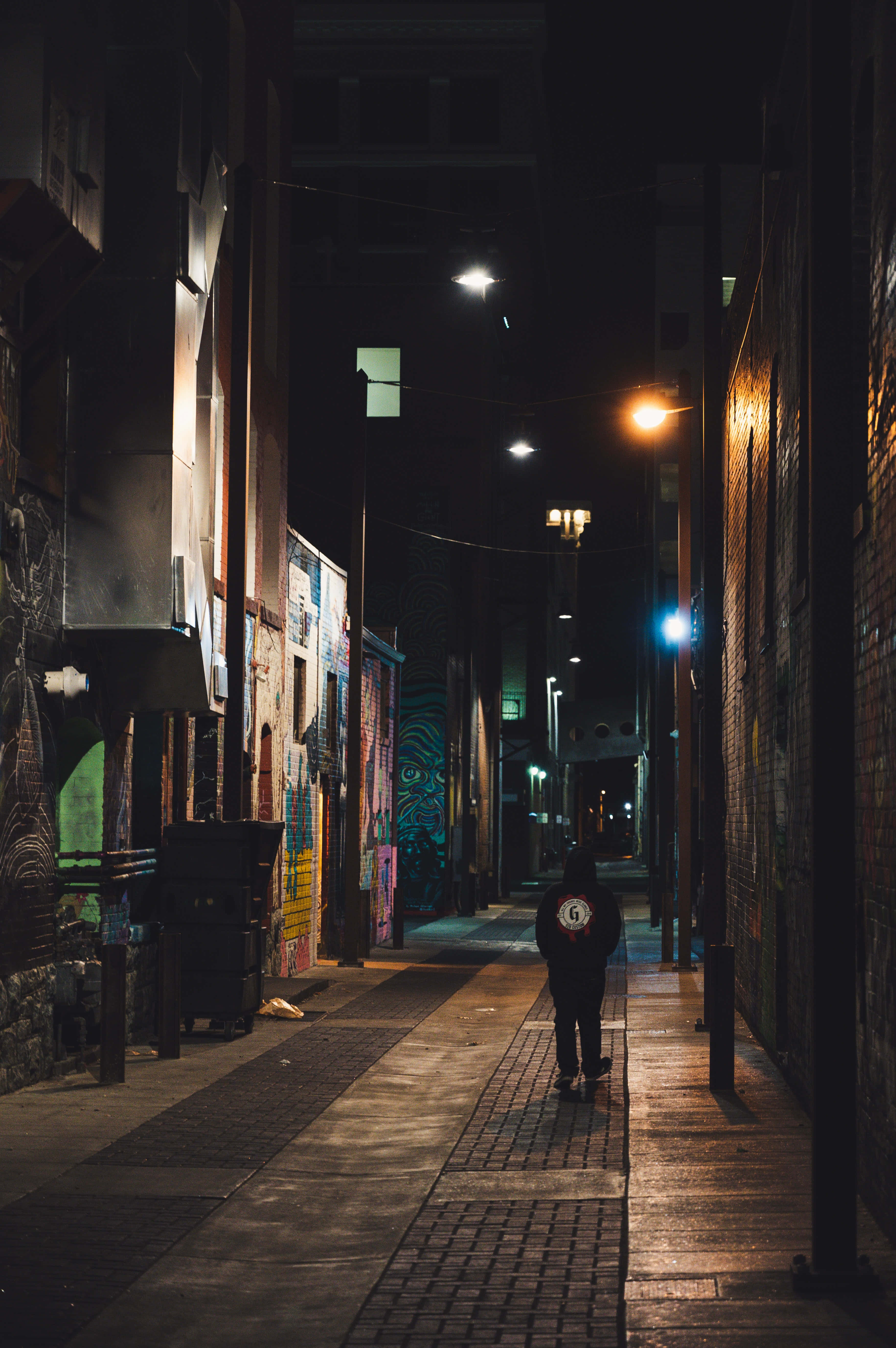 Dark Alleyway 桌布 壁紙 — 一個孤獨的身影在夜晚沿著狹窄的小巷行走，被街燈和磚牆上色彩繽紛的塗鴉照亮。