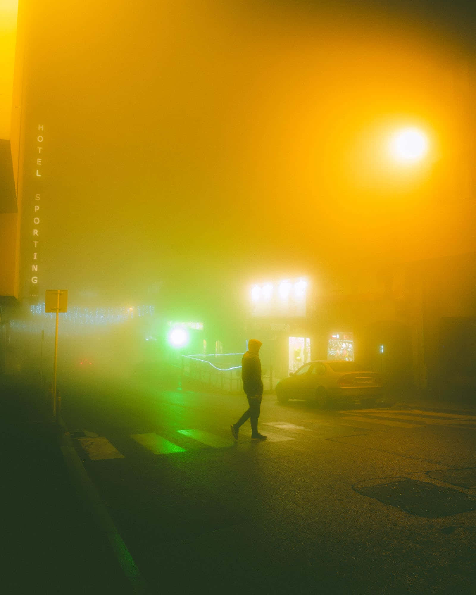 San Francisco Fog 桌布 壁紙 — 一個孤獨的身影在夜晚穿過霧氣瀰漫的街道，被酒店和商店櫥窗詭異的黃色和綠色霓虹燈照亮。
