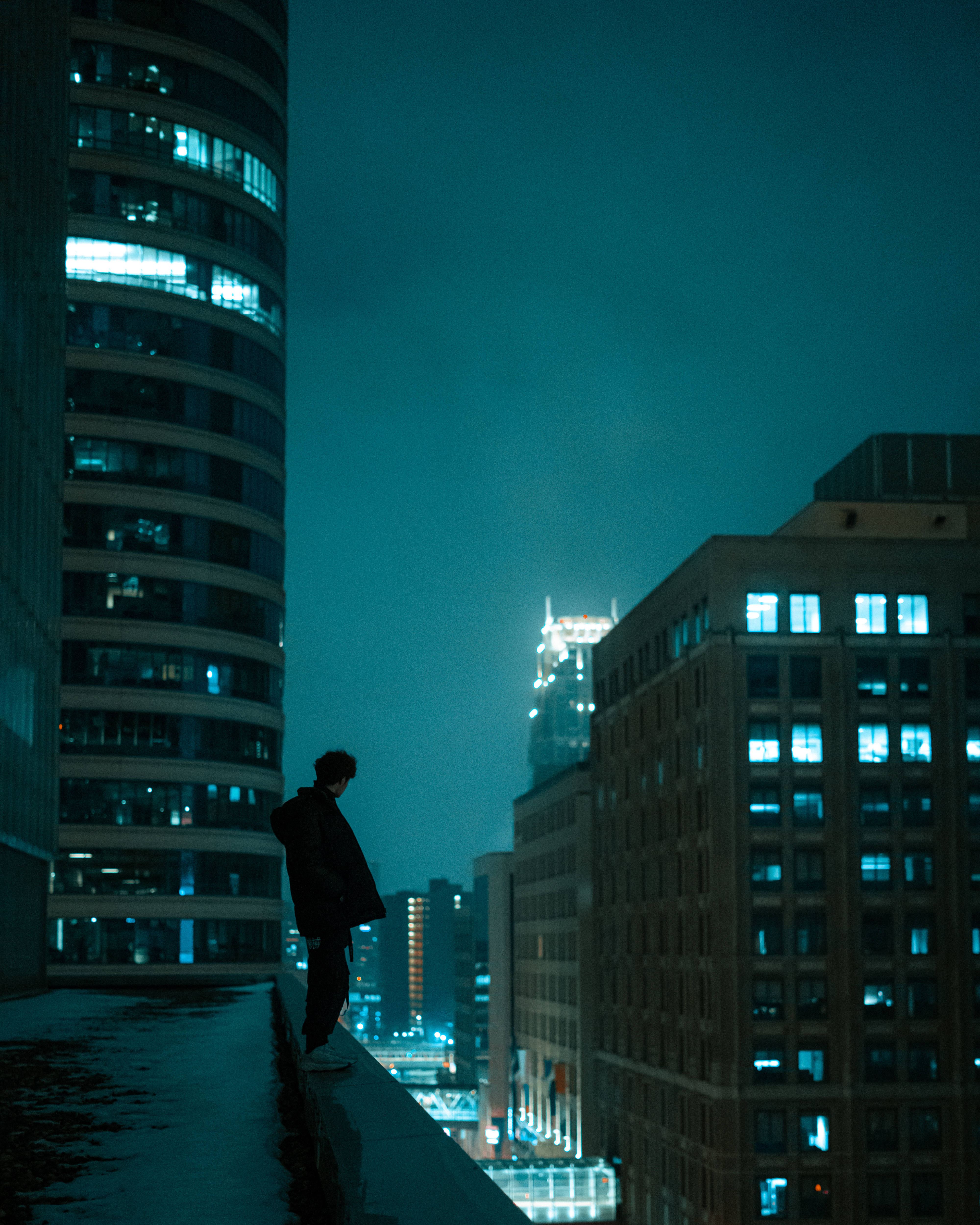 青色GIF 桌布 壁紙 — 一個孤獨的身影在夜晚站在屋頂上，俯瞰著城市景觀，窗戶閃爍著光芒，天空呈現深藍綠色。
