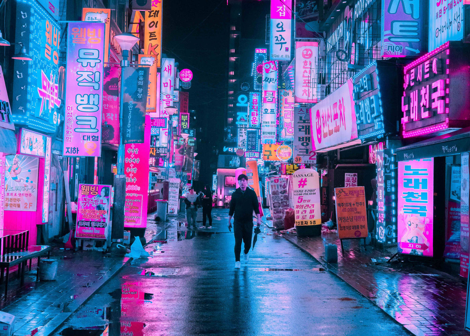 一個人在夜晚走過霓虹燈照亮的韓國街道，潮濕路面有倒影。 — 東京 Ravens 壁紙