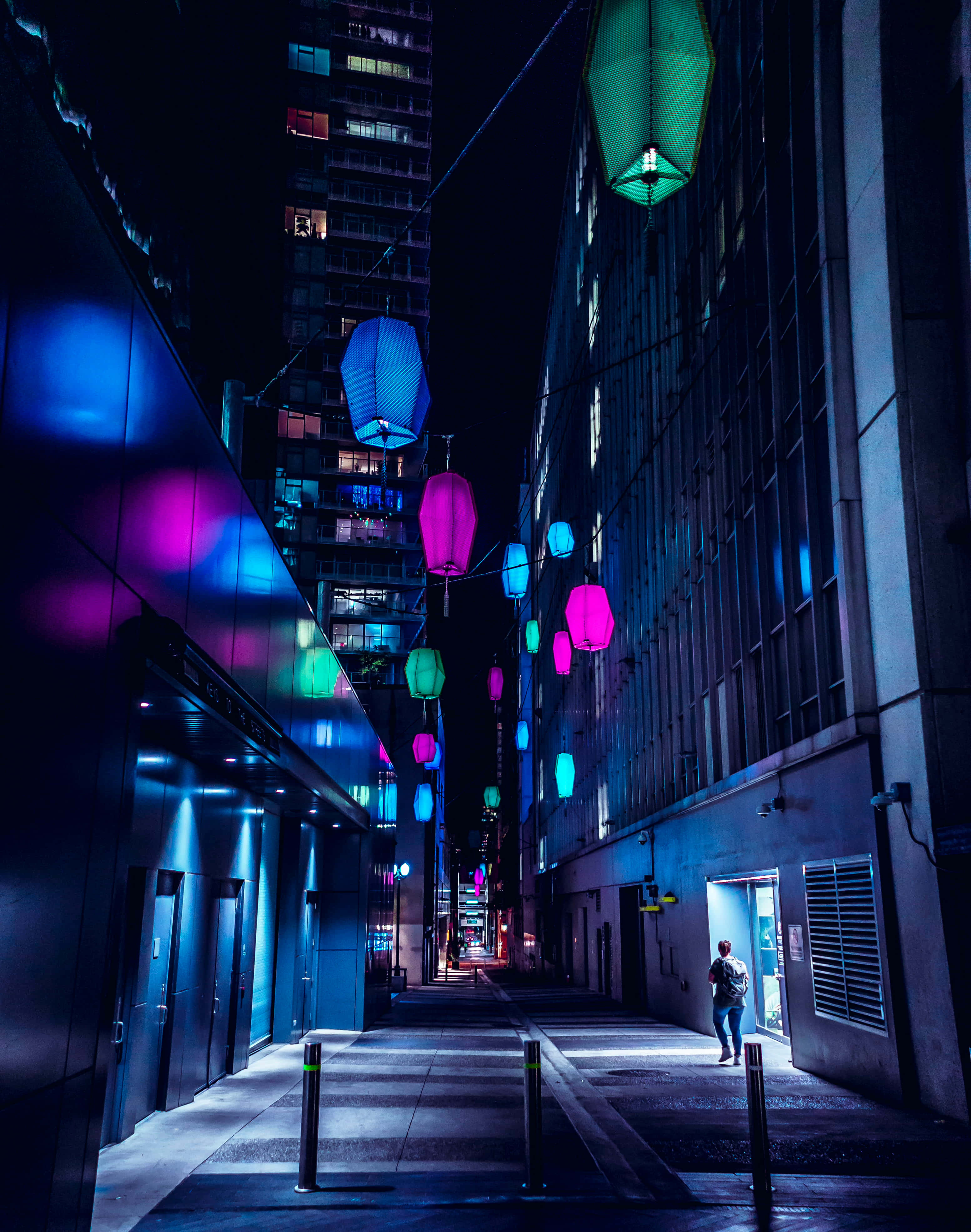 City Lights 桌布 壁紙 — 一個孤獨的身影在夜晚走在一條霓虹燈照亮嘅城市小巷，周圍係掛喺高樓之間嘅發光粉紅色同藍色燈籠。