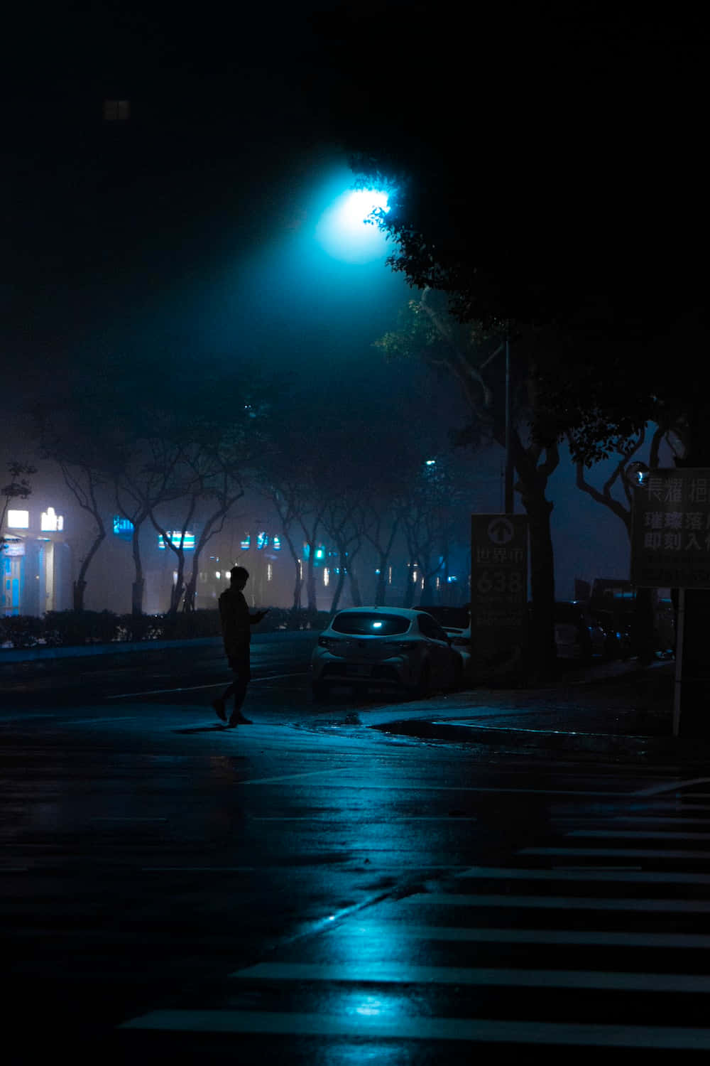 Dark Crossroad 桌布 壁紙 — 一個孤獨的身影在夜晚走過濕漉漉的城市街道，頭頂是發出藍光的街燈，投下長長的影子。