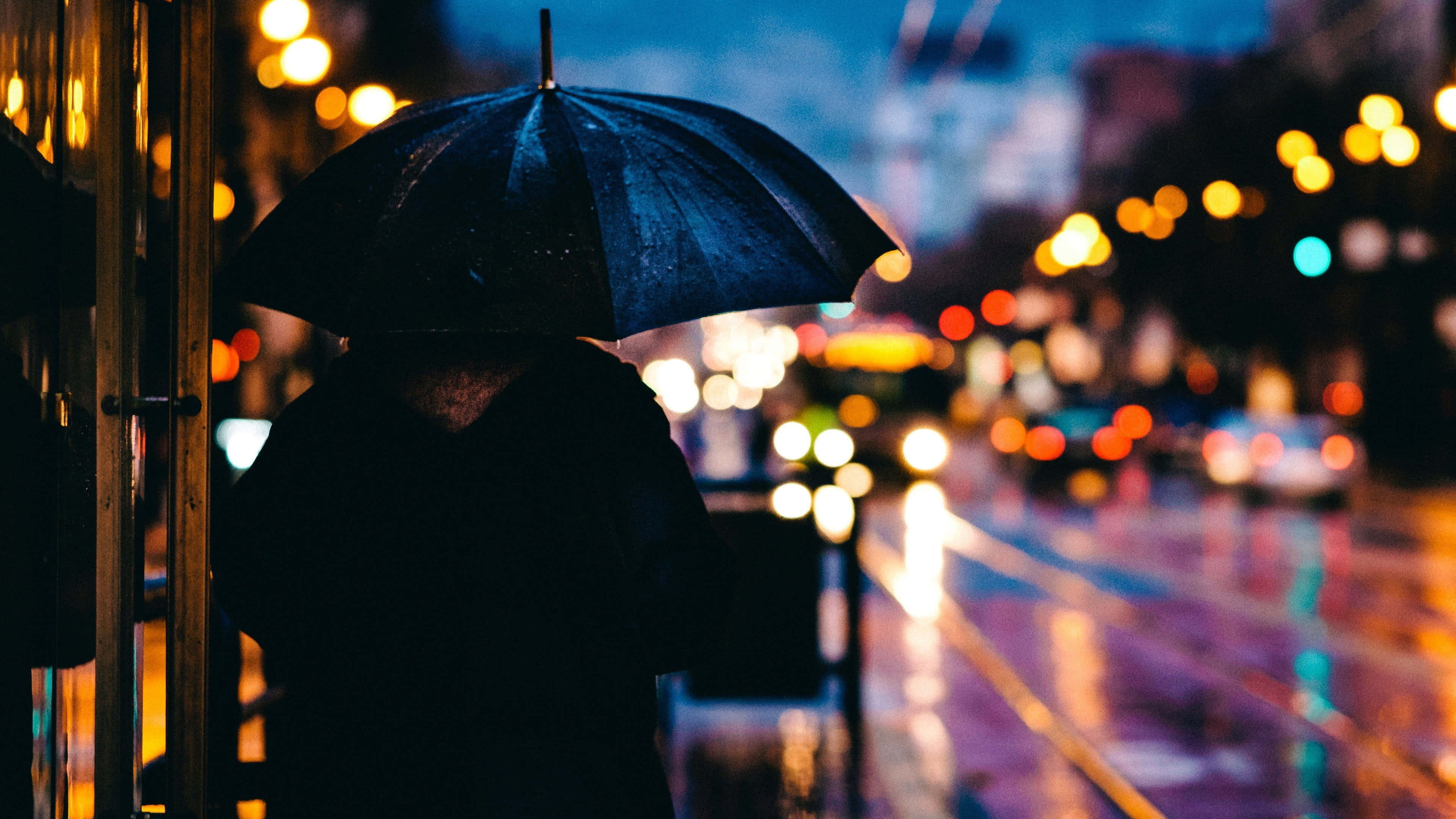 下雨 桌布 壁紙 — 一個孤獨的身影在夜晚霓虹燈照亮的城市街道上撐著黑色雨傘擋雨。