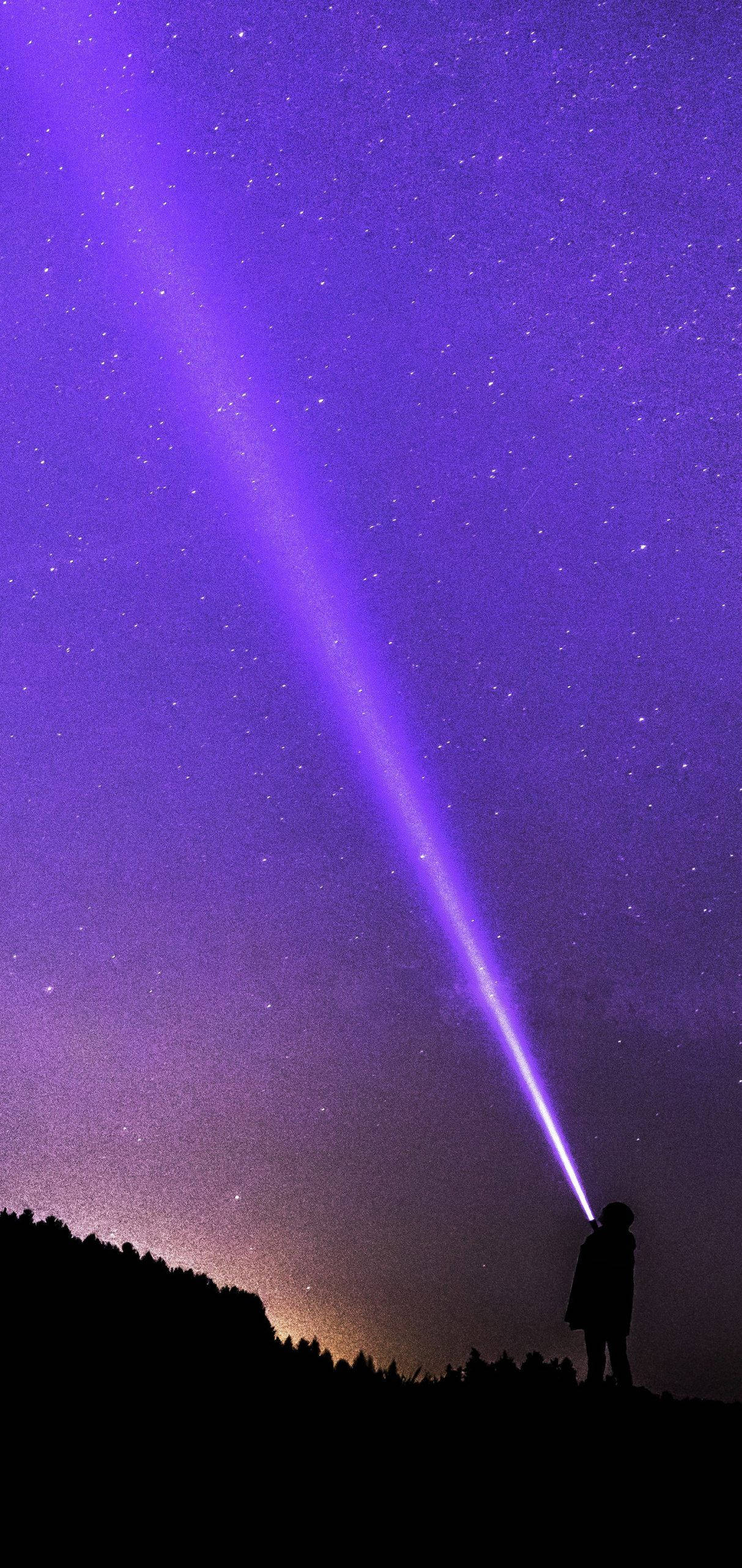  壁紙 — 一個孤獨的身影在夜間站在山丘上，將一道明亮的紫色光束射向繁星點點的紫羅蘭色天空。