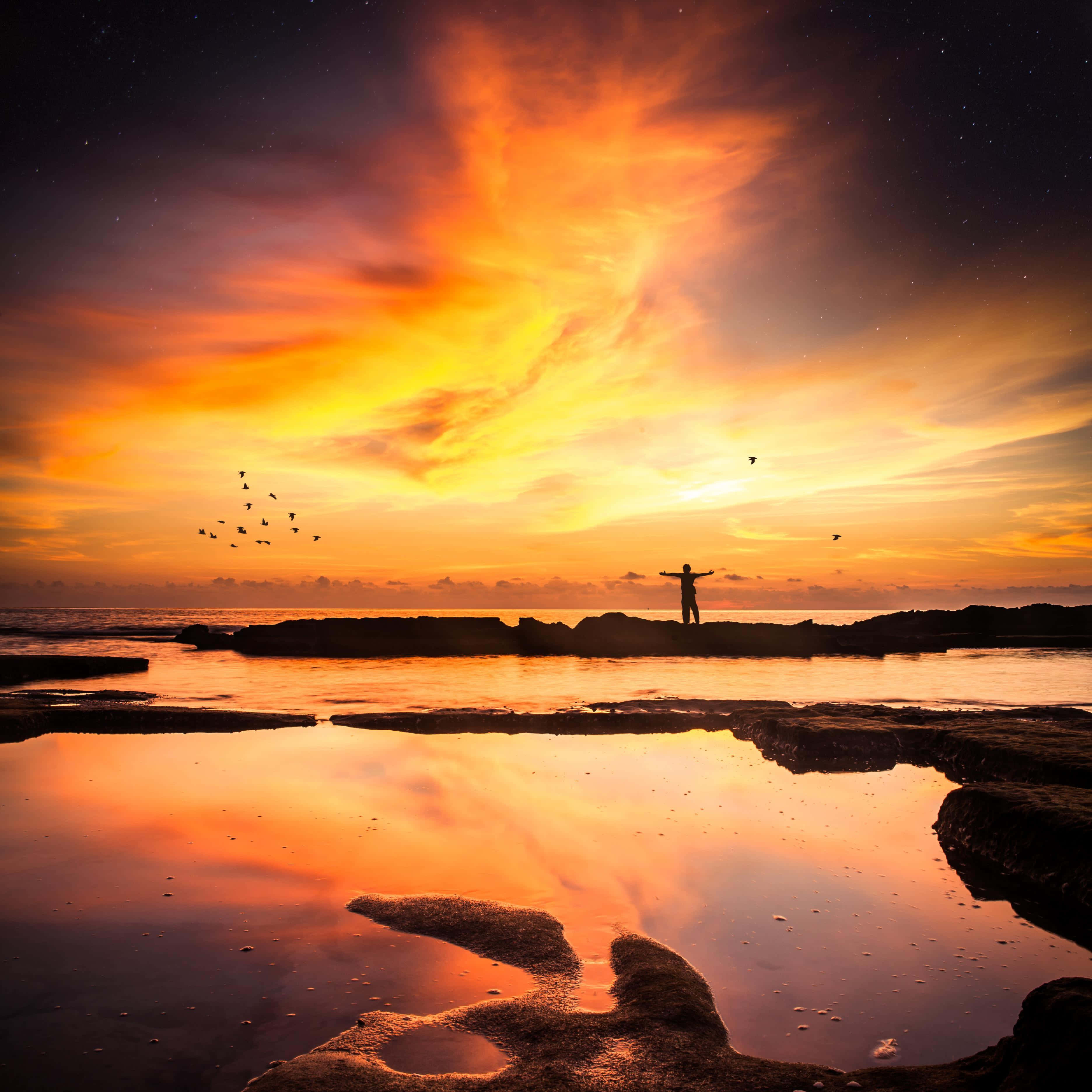 Sunset Reflection 桌布 壁紙 — 一個孤獨的身影在岩石海岸上張開雙臂，熾熱的日落雲彩在星空下的潮汐池中倒映。