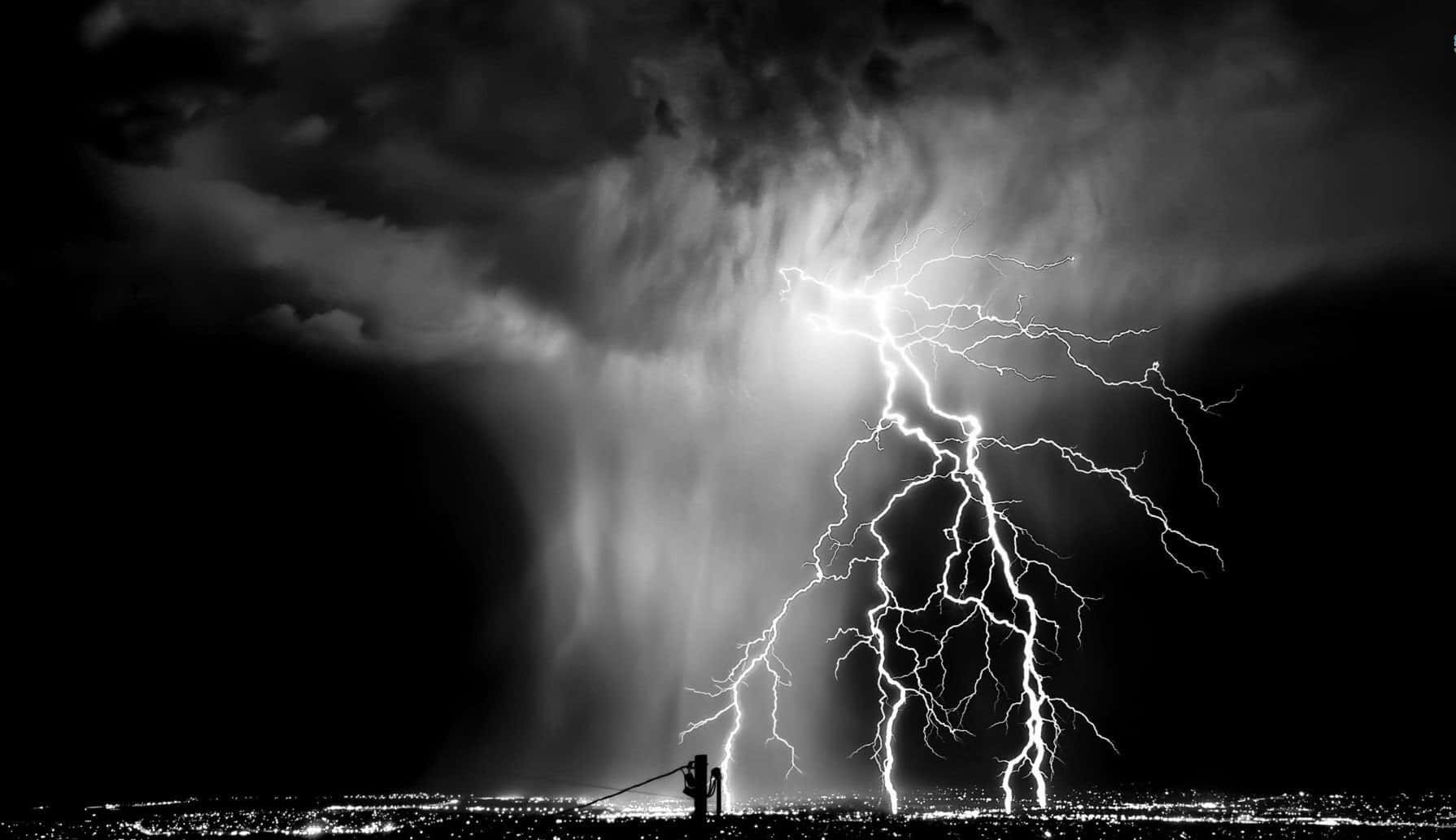 Black And White Storm 桌布 壁紙 — 一個孤獨的身影在戲劇性的夜空下呈現剪影，巨大的閃電照亮了遠處城市景觀上方的暴風雲。