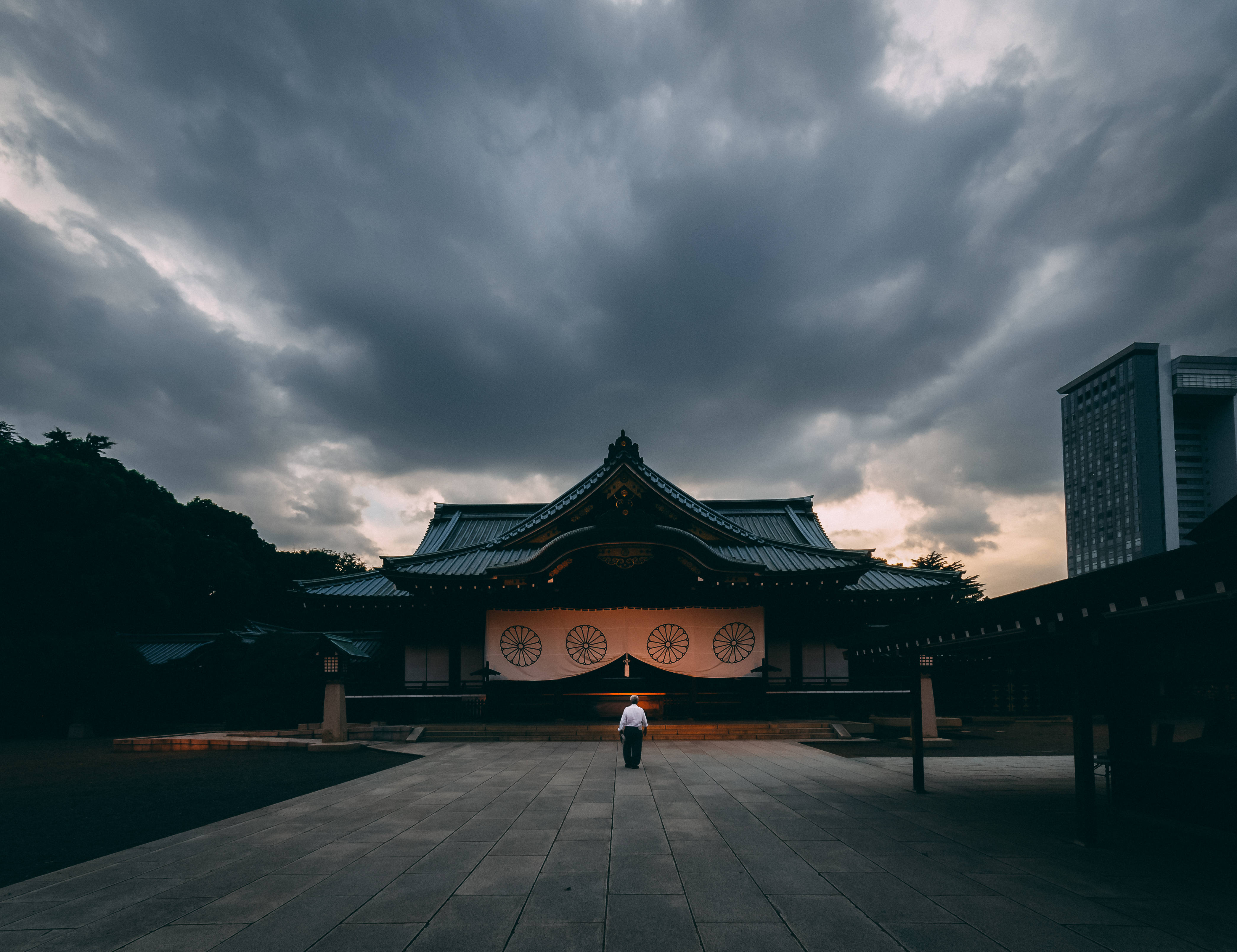 日本 桌布 壁紙 — 一個孤獨的身影在戲劇性的暴風雨天空下走向傳統日本神社，背景有現代建築。