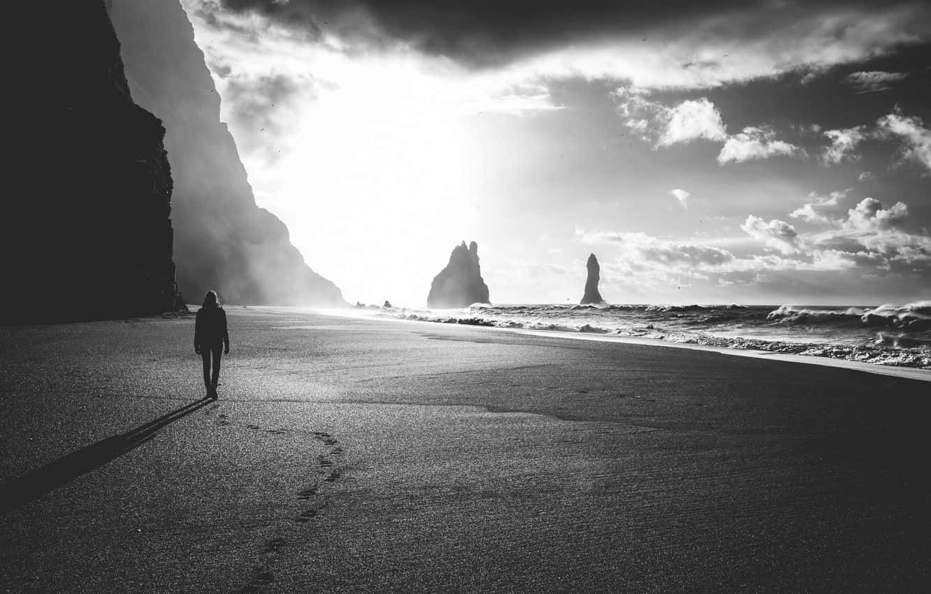 Black And White Beach 桌布 壁紙 — 一個孤獨的身影在明亮的天空下，沿著一個戲劇性的黑白海灘行走，海灘上有高聳的懸崖和海蝕柱。