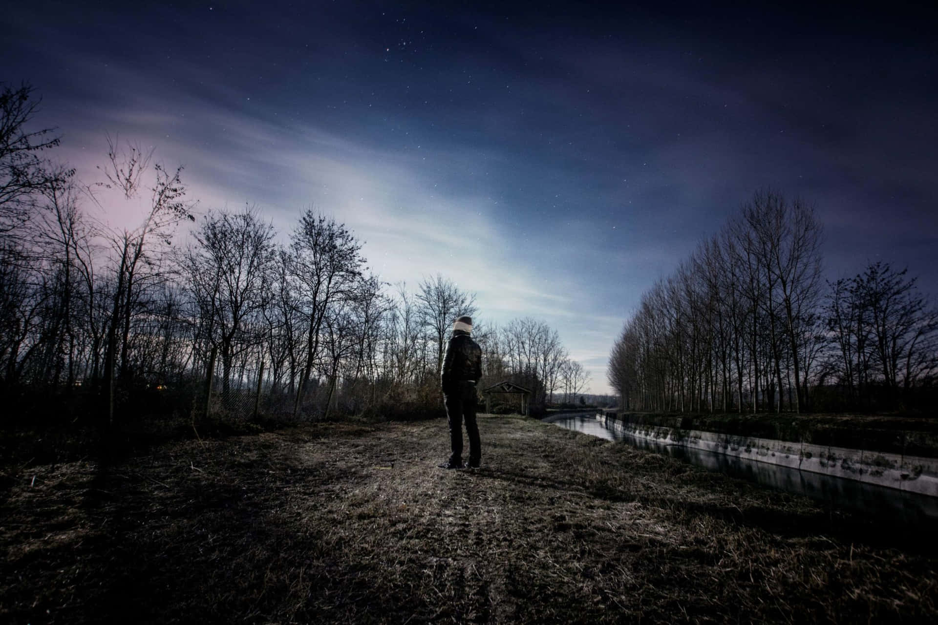 Alone Boy 桌布 壁紙 — 一個孤獨的身影在暮色中站在運河旁，被光禿禿的樹木框住，天空繁星點點，帶有柔和的紫色雲彩。