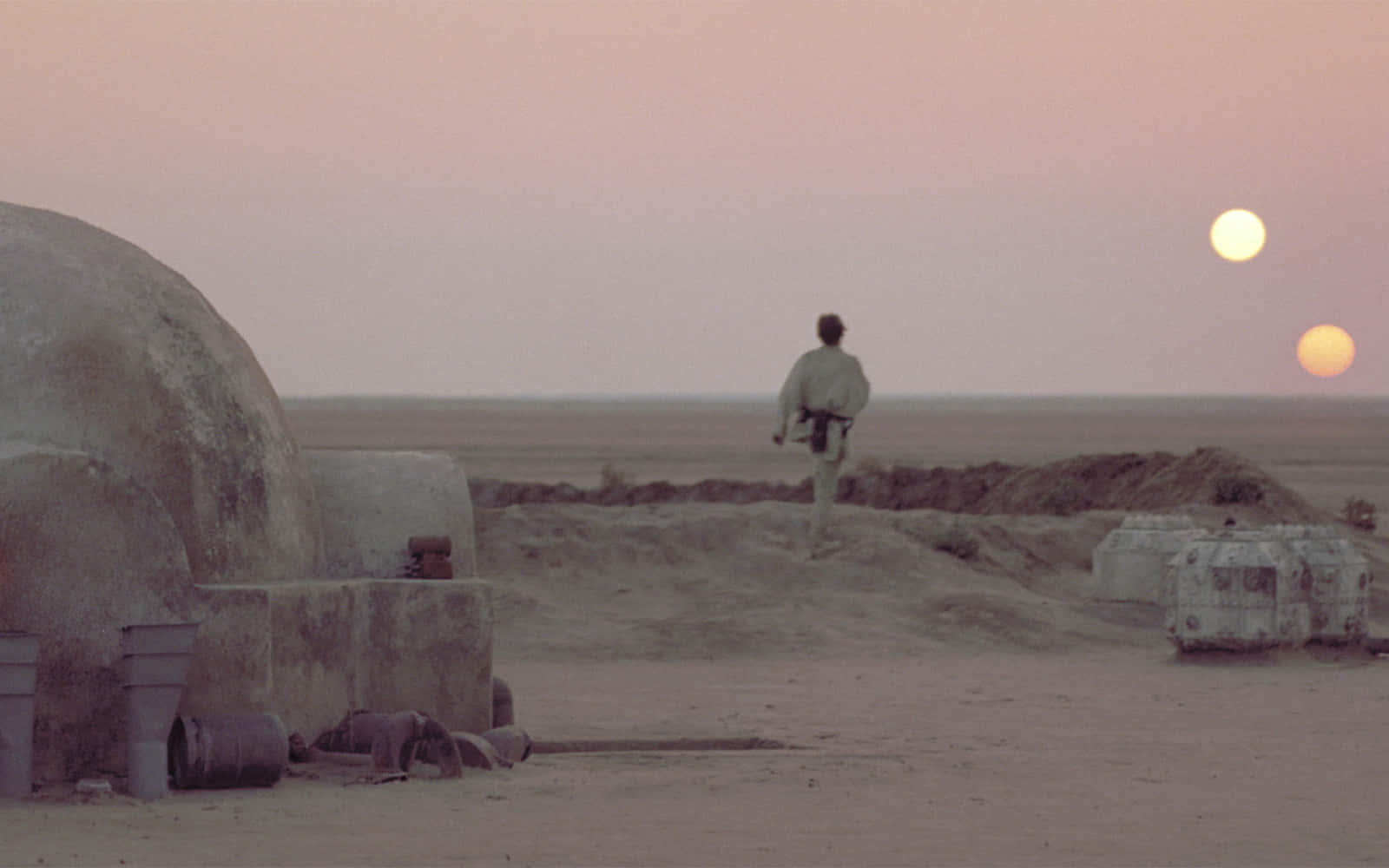 Jedha 桌布 壁紙 — 一個孤獨的身影在暮色中走過沙漠景觀，經過圓頂狀的結構，粉紅色的天空中閃耀著兩個太陽。