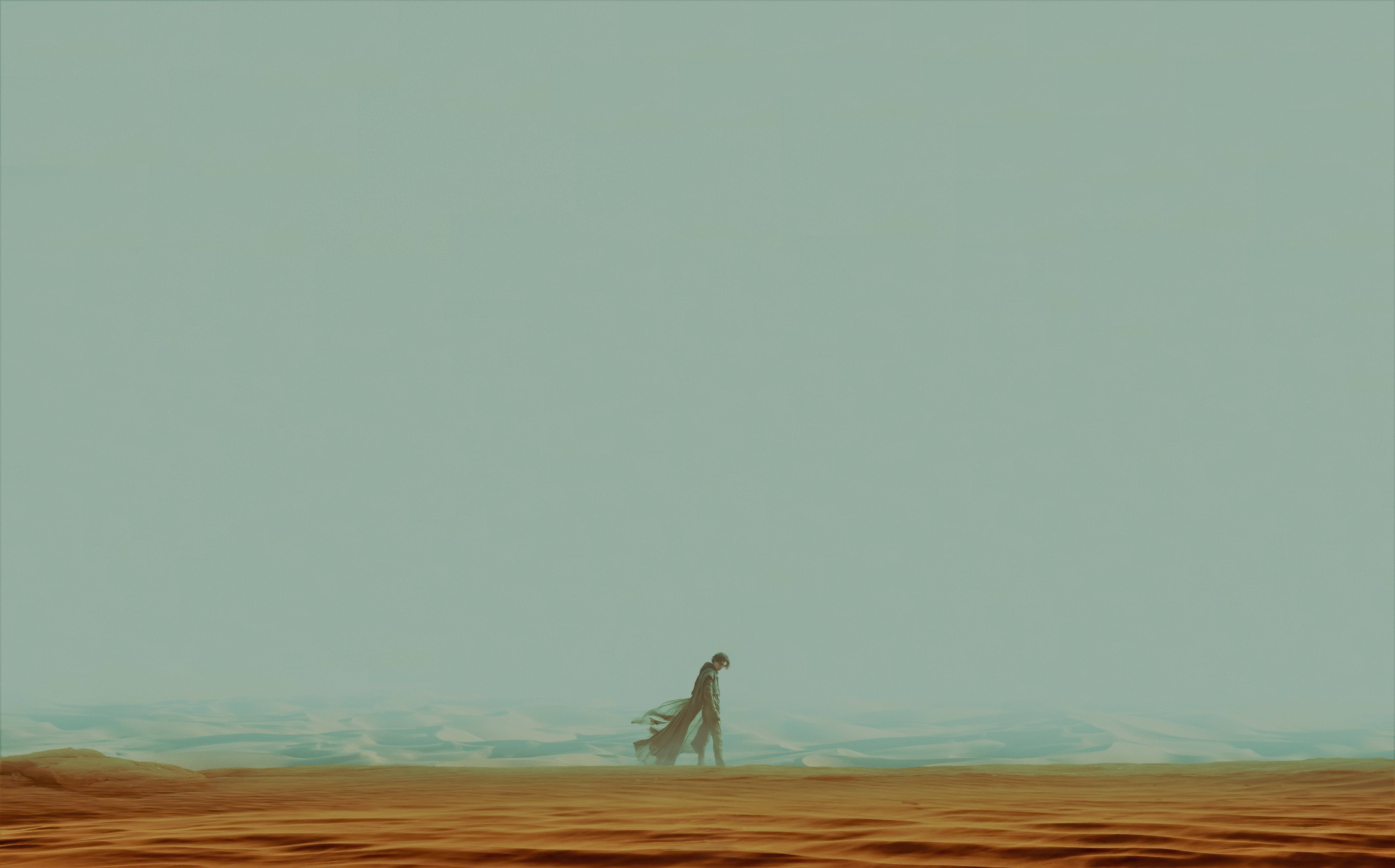 Dune 2021 桌布 壁紙 — 一個孤獨的身影在朦朧的青綠色天空下，穿越廣闊的沙漠，連綿的沙丘一直延伸到地平線。