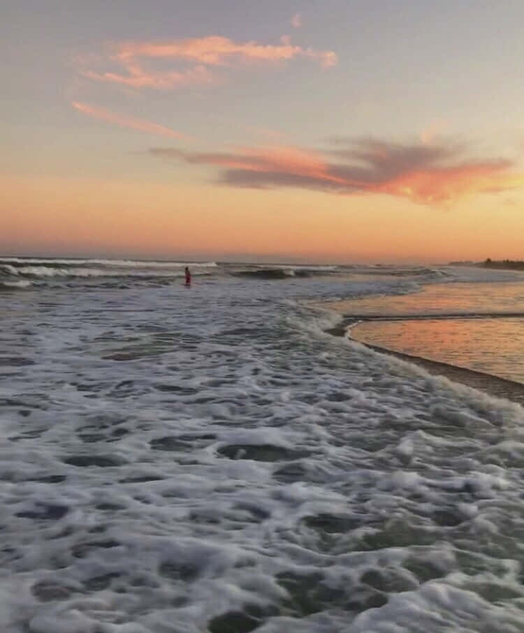 Beach Aesthetic 桌布 壁紙 — 一個孤獨的身影在海洋波浪中涉水而行，粉紅色和橙色的雲彩在日落時分在地平線上發光。